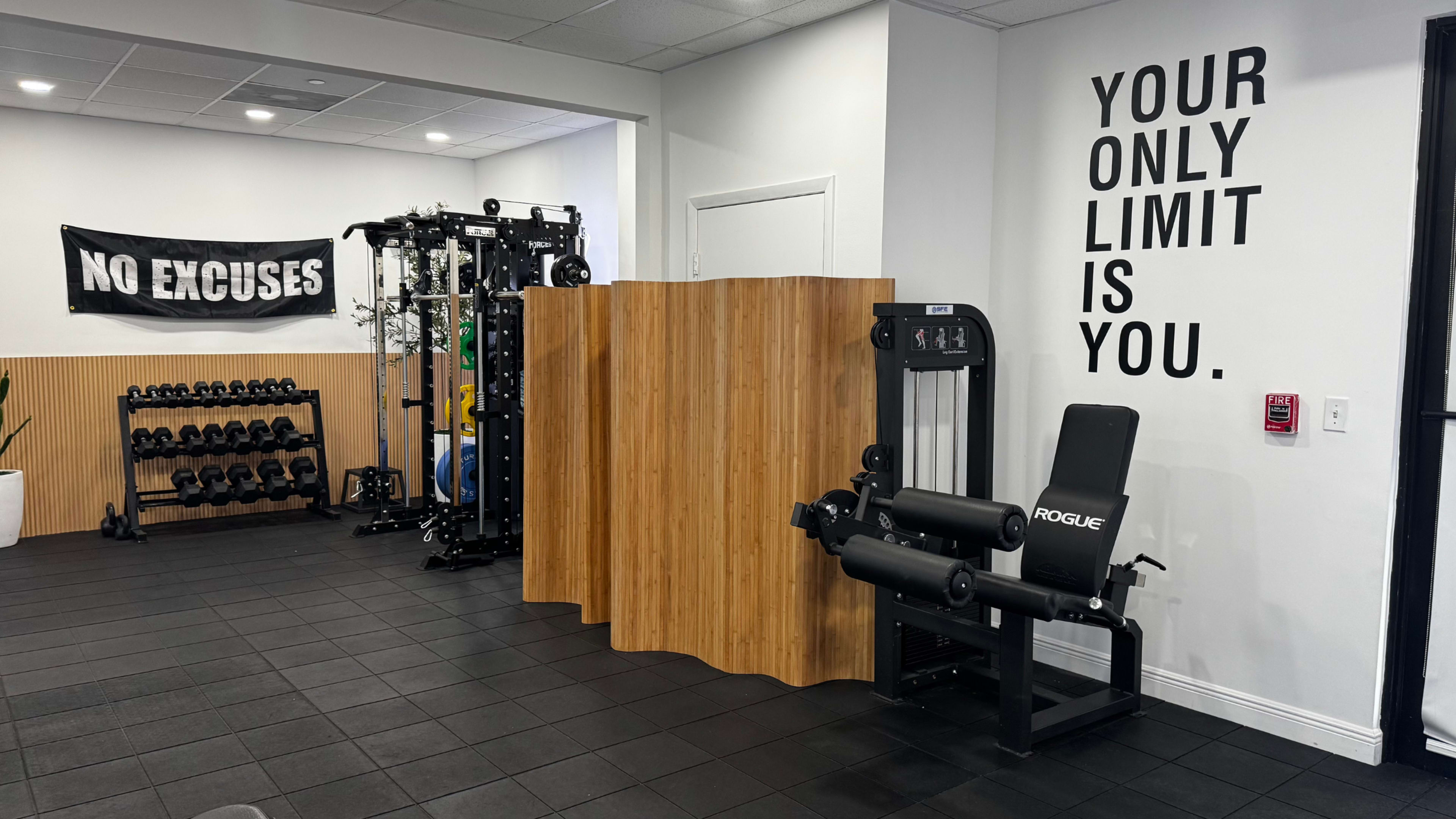 The image shows a gym interior featuring weightlifting equipment, a wooden partition, and motivational signage on the wall.