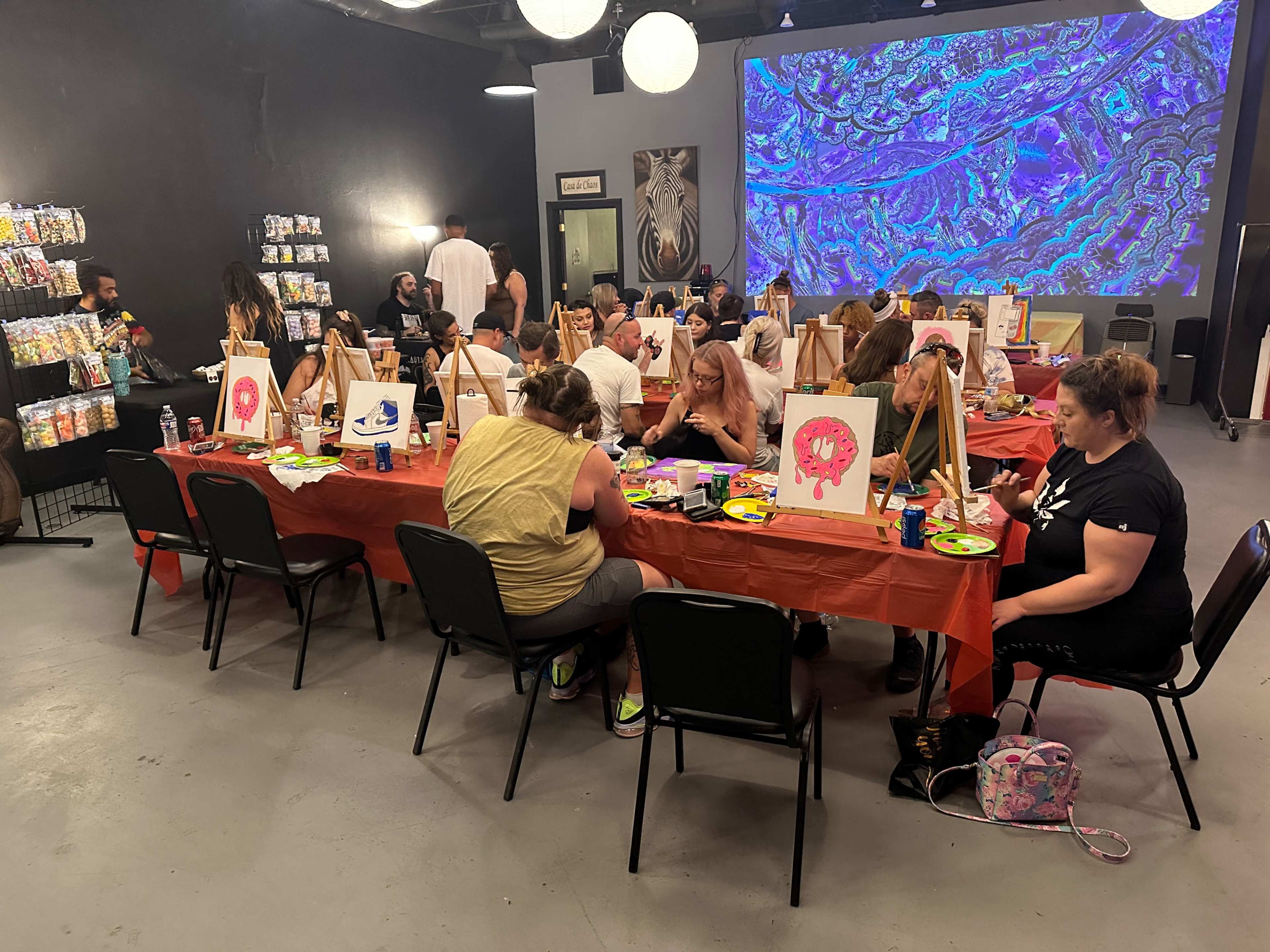 A group of people are sitting at tables in a semi-lit room, engaged in a painting activity while snacks and colorful canvases are displayed.