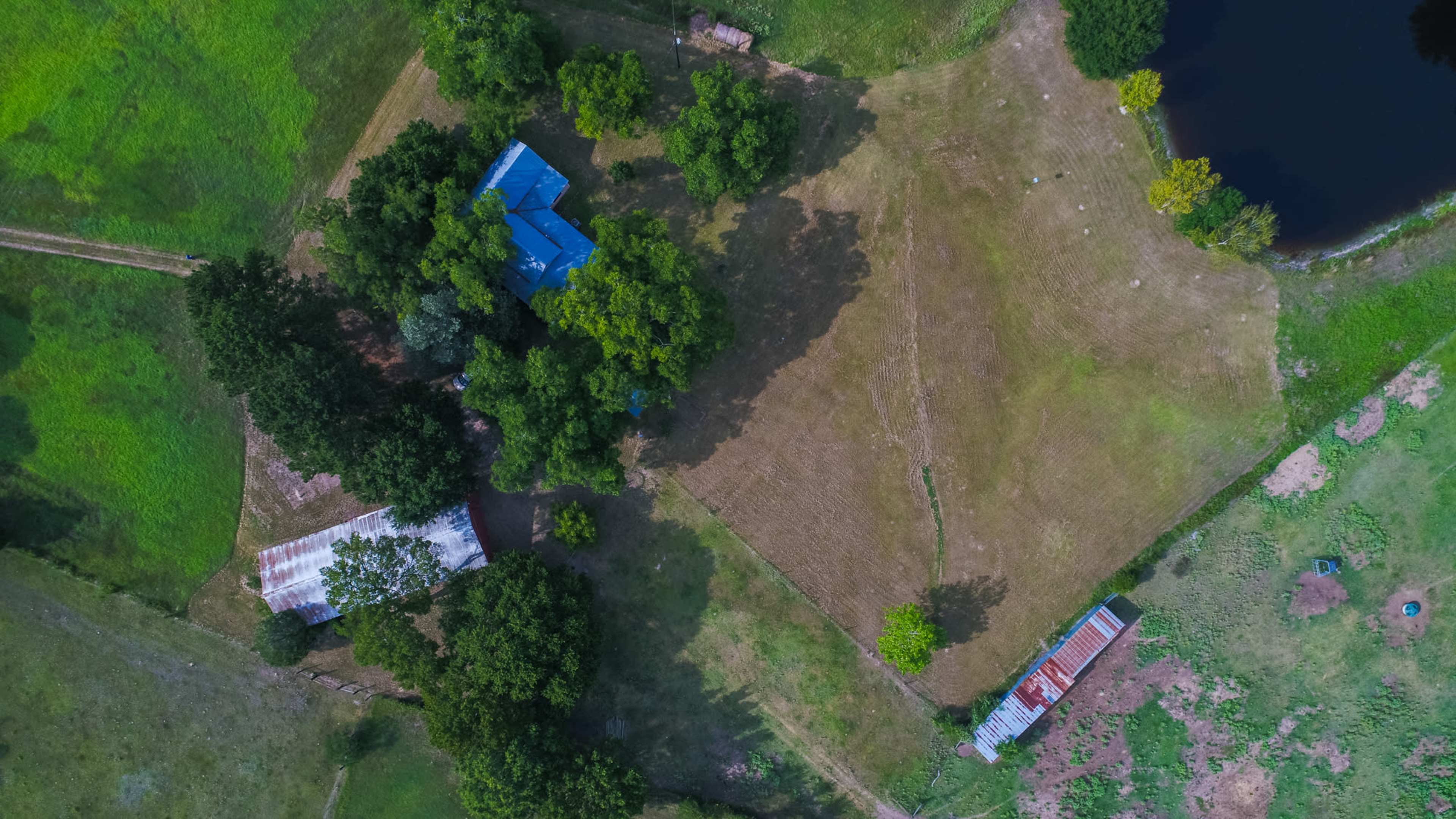 An aerial view shows a rural property featuring a blue-roofed house, a metal shed, and a pond surrounded by trees and open fields.