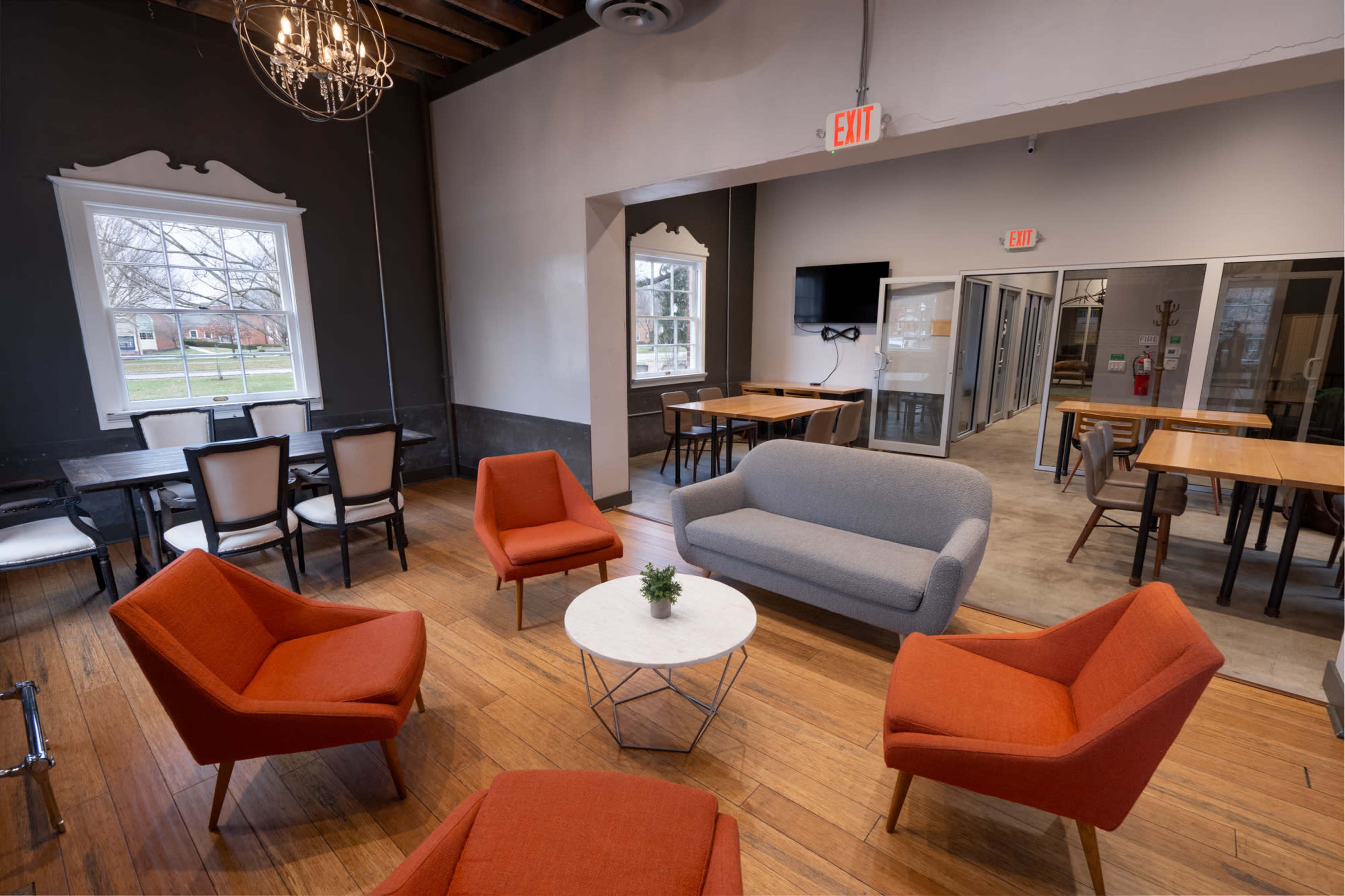 The image shows a modern, open-concept lounge area with orange chairs and a gray sofa arranged around a white coffee table, complemented by wooden floors and large windows.