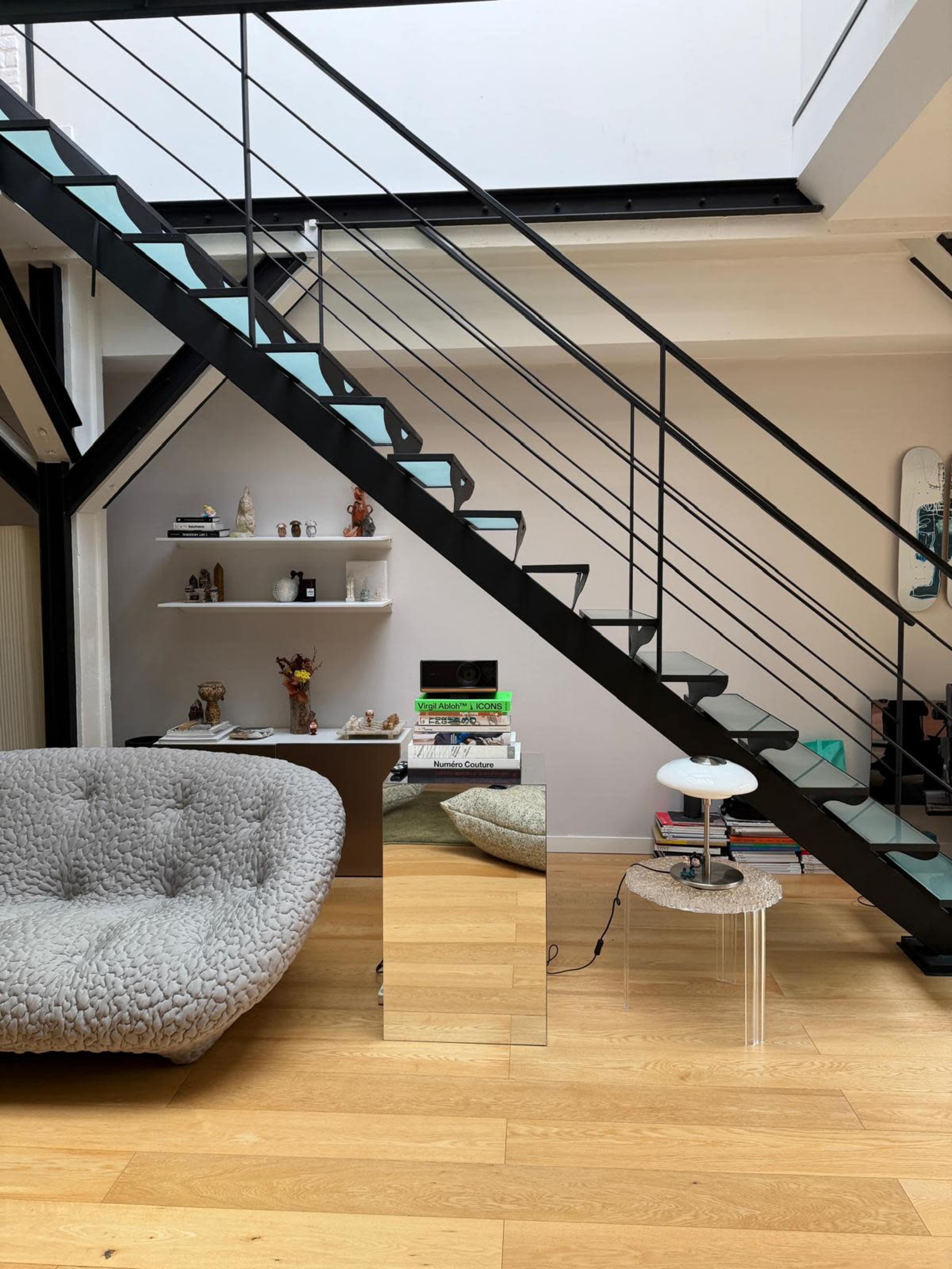 The image shows a modern living space with a gray tufted sofa, a mirrored side table, and a staircase with black metal railings and light blue steps.