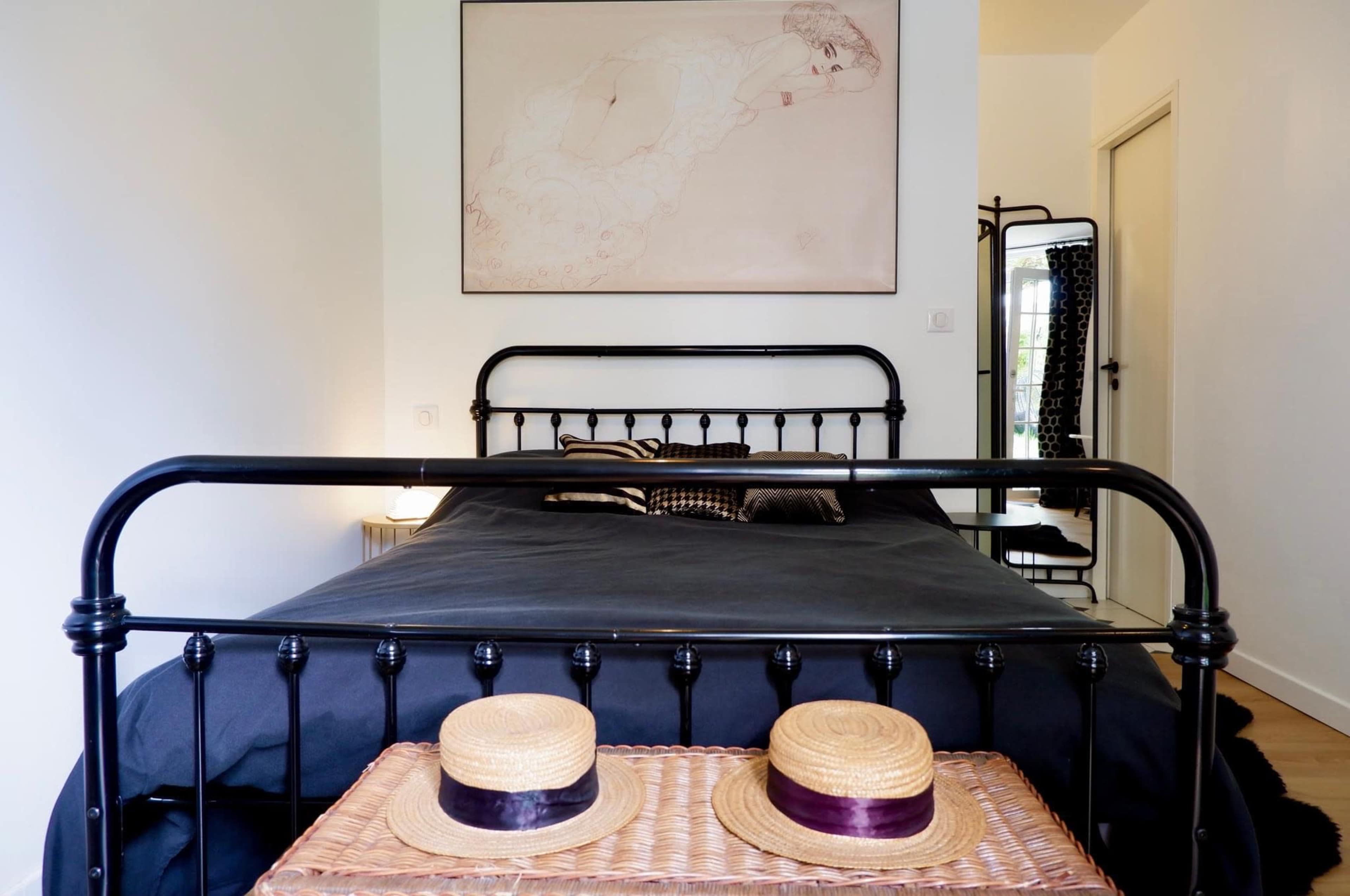 The image shows a simple bedroom featuring a black metal bed, a large framed artwork on the wall, and two straw hats placed on a woven basket at the foot of the bed.