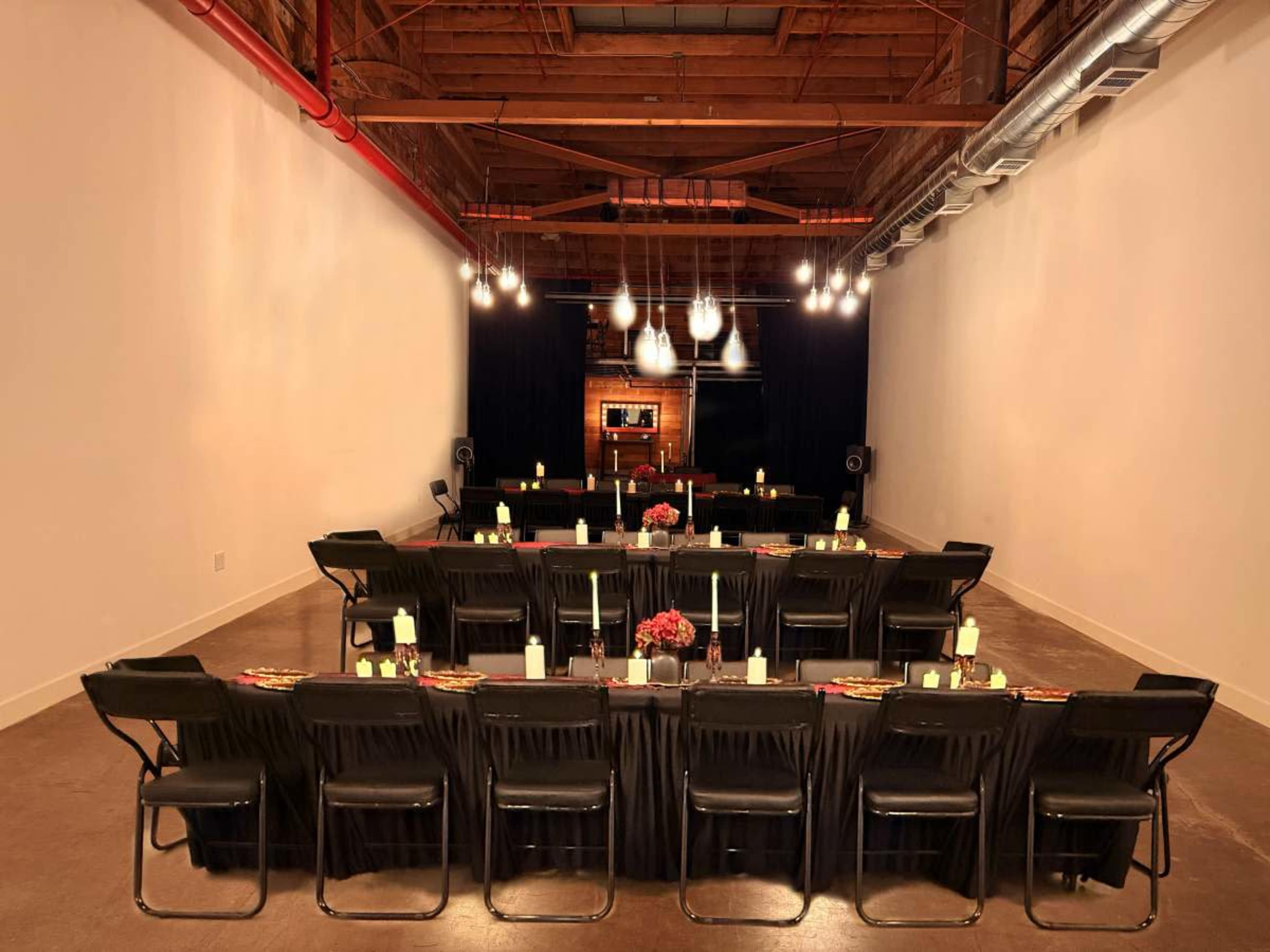 The image shows a spacious event room set up with rows of black chairs and tables, elegantly arranged with candles and floral centerpieces, facing a stage area with lighting.