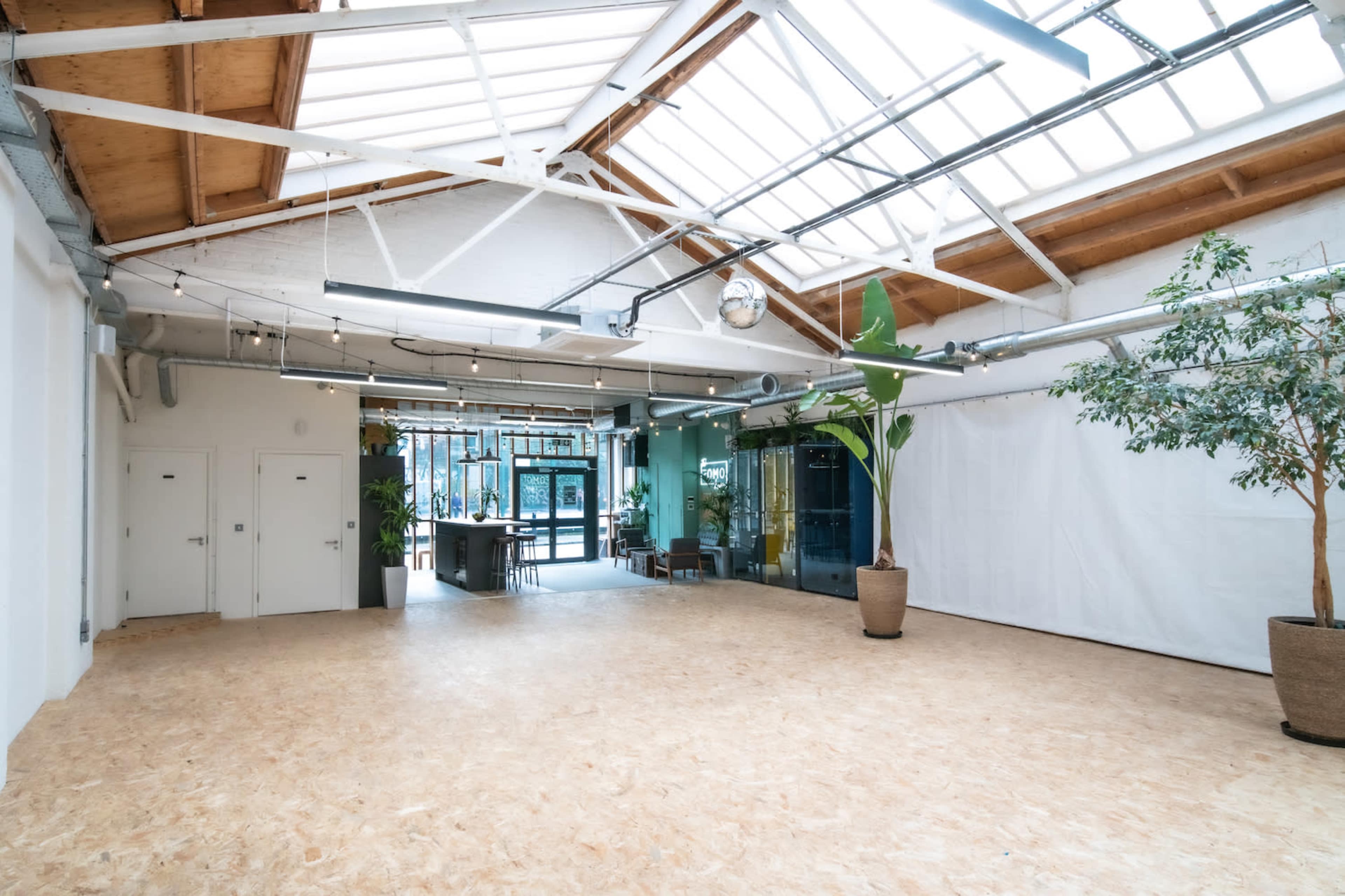 A spacious, well-lit interior with a wooden floor, large windows, and potted plants, featuring a seating area in the background.