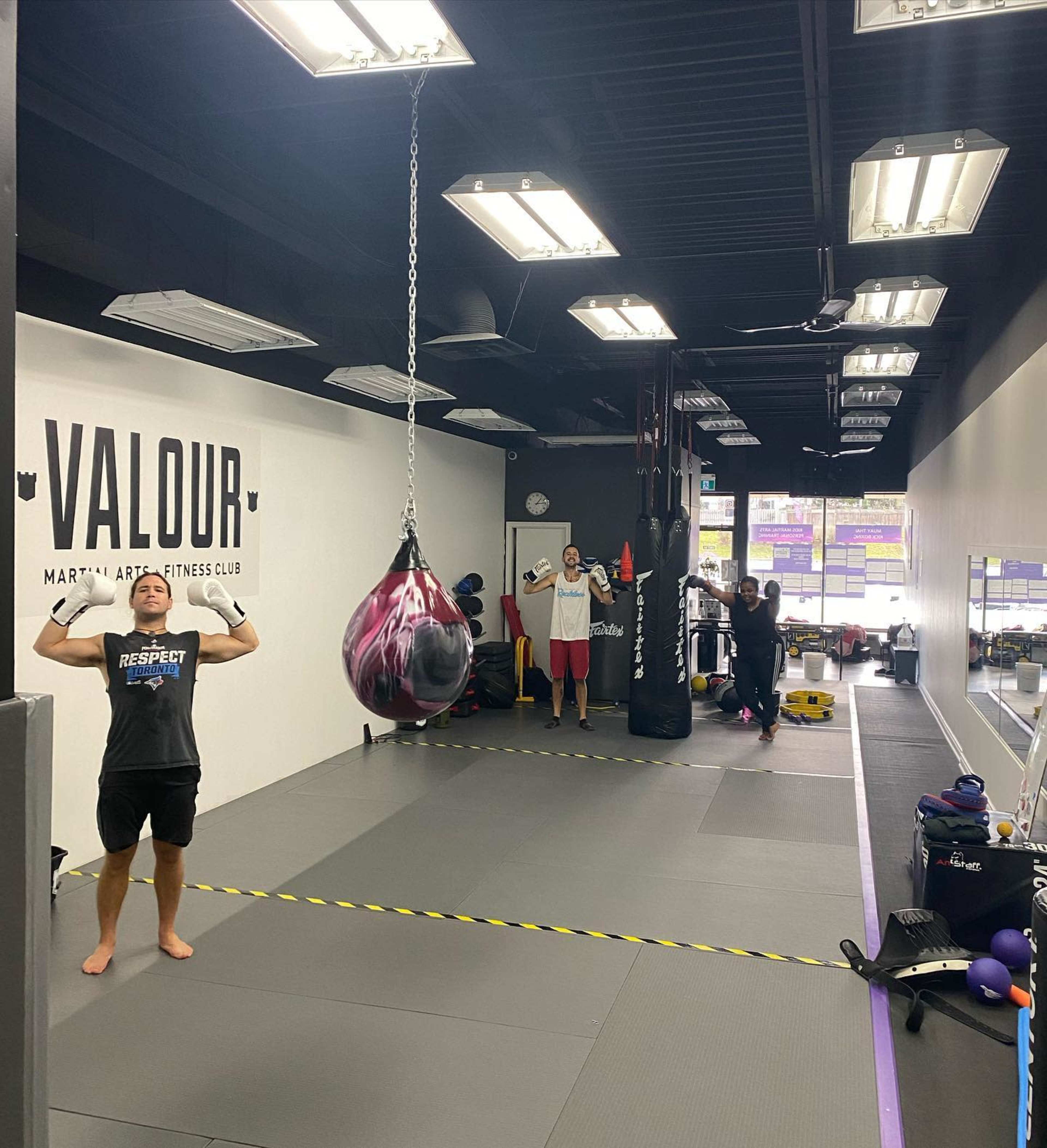 A fitness club interior features a training area with a heavy bag hanging, individuals exercising, and equipment displayed along the walls.