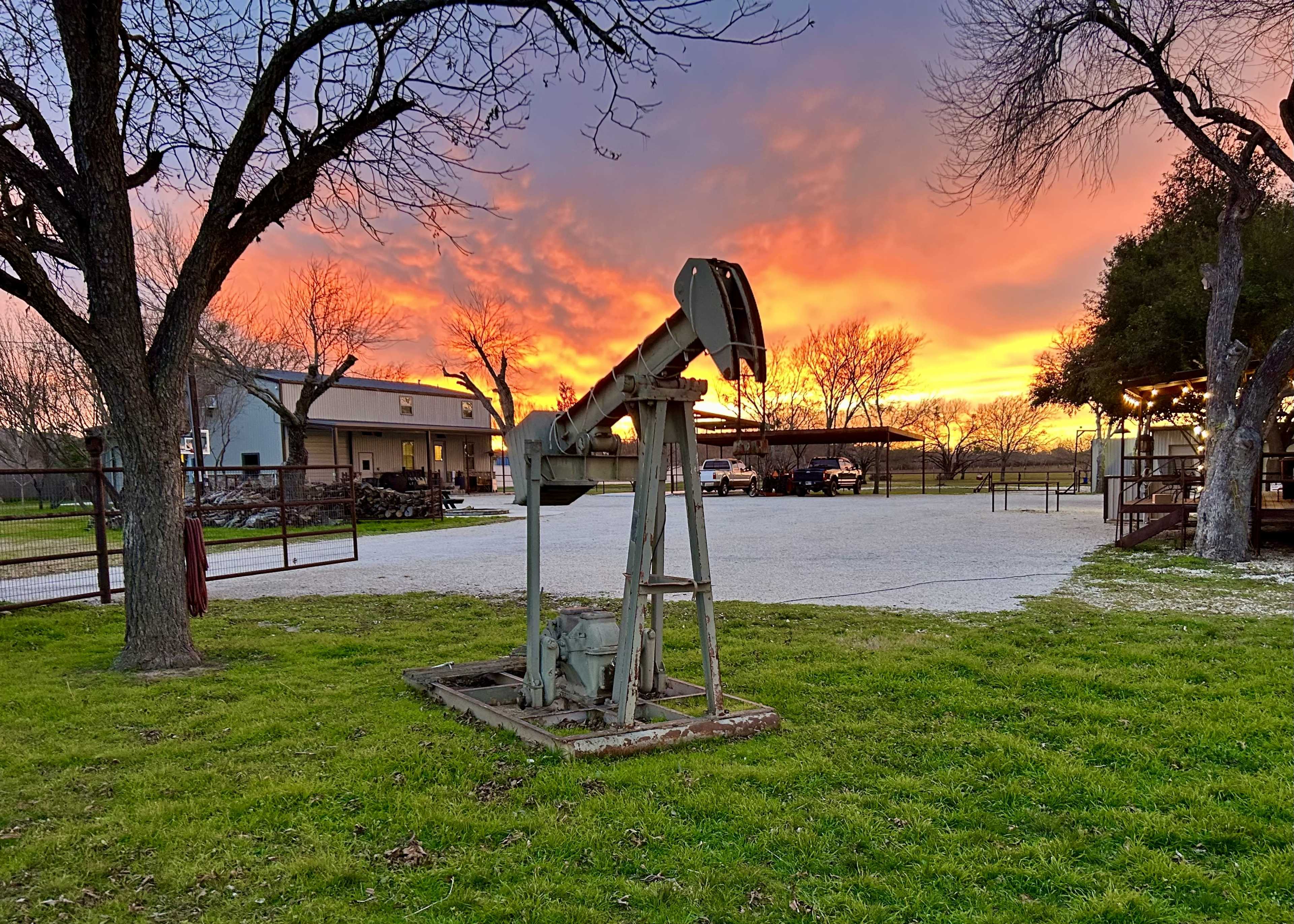 A vintage oil pump jack stands in the foreground of a rural property against a vibrant sunset sky.