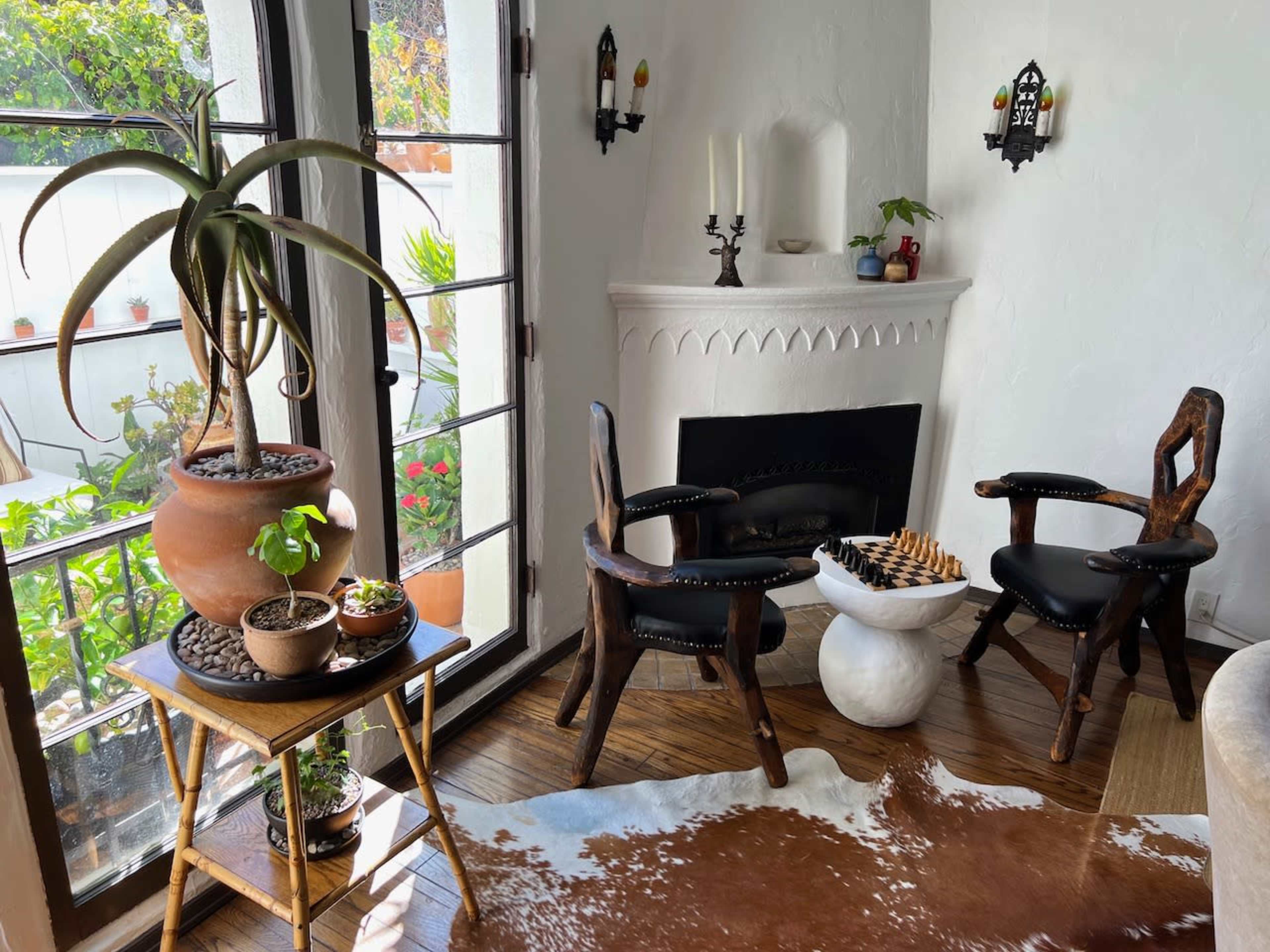 A cozy nook features two wooden chairs, a marble table with a chess set, and a large potted plant, all arranged near a fireplace and a window overlooking a garden.