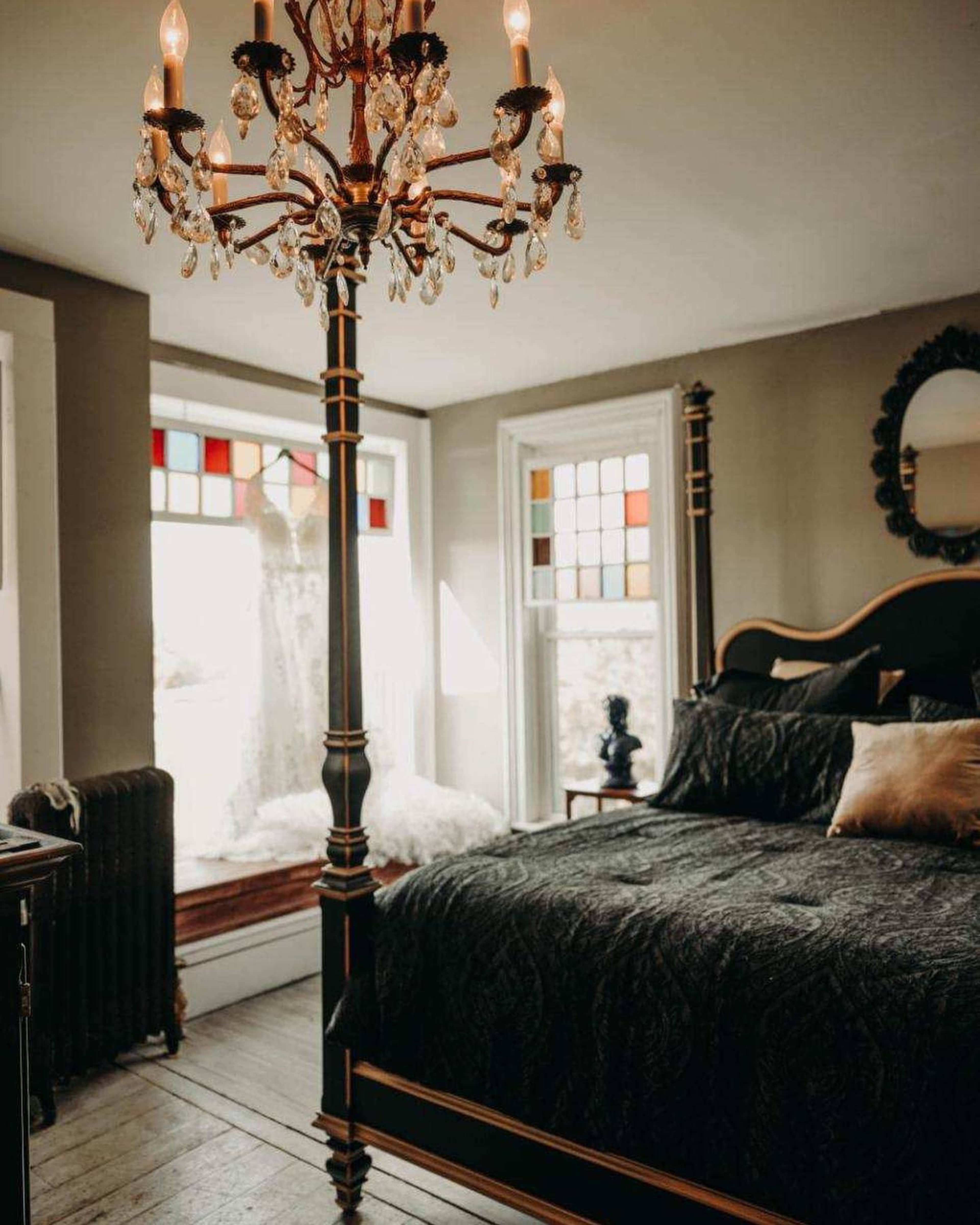 A bedroom featuring a chandelier, a dark bedspread, and a window with stained glass alongside a white dress draped on a nearby window seat.