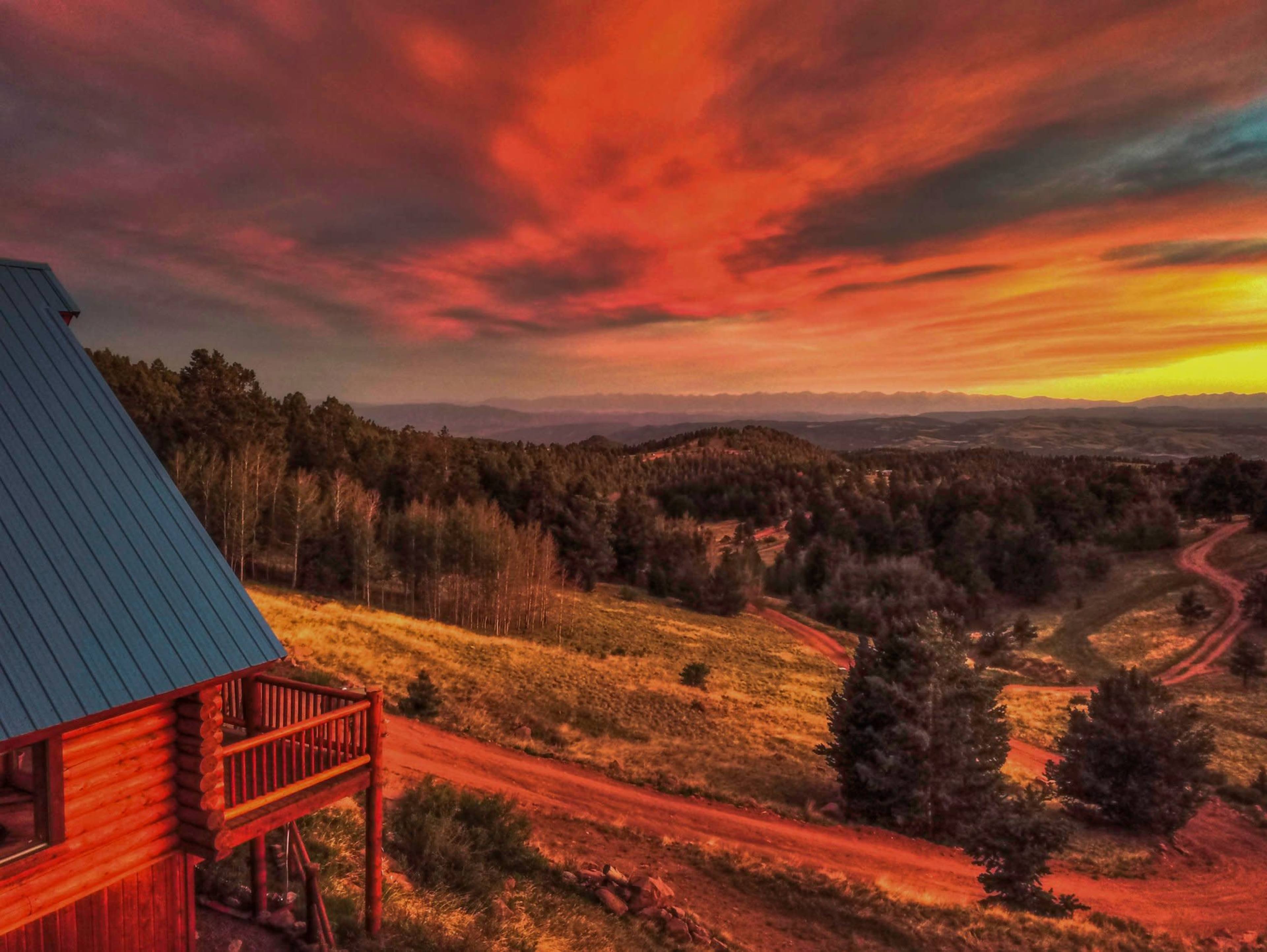 A cabin with a blue roof overlooks a winding dirt road leading through rolling hills at sunset, casting vibrant colors across the sky.