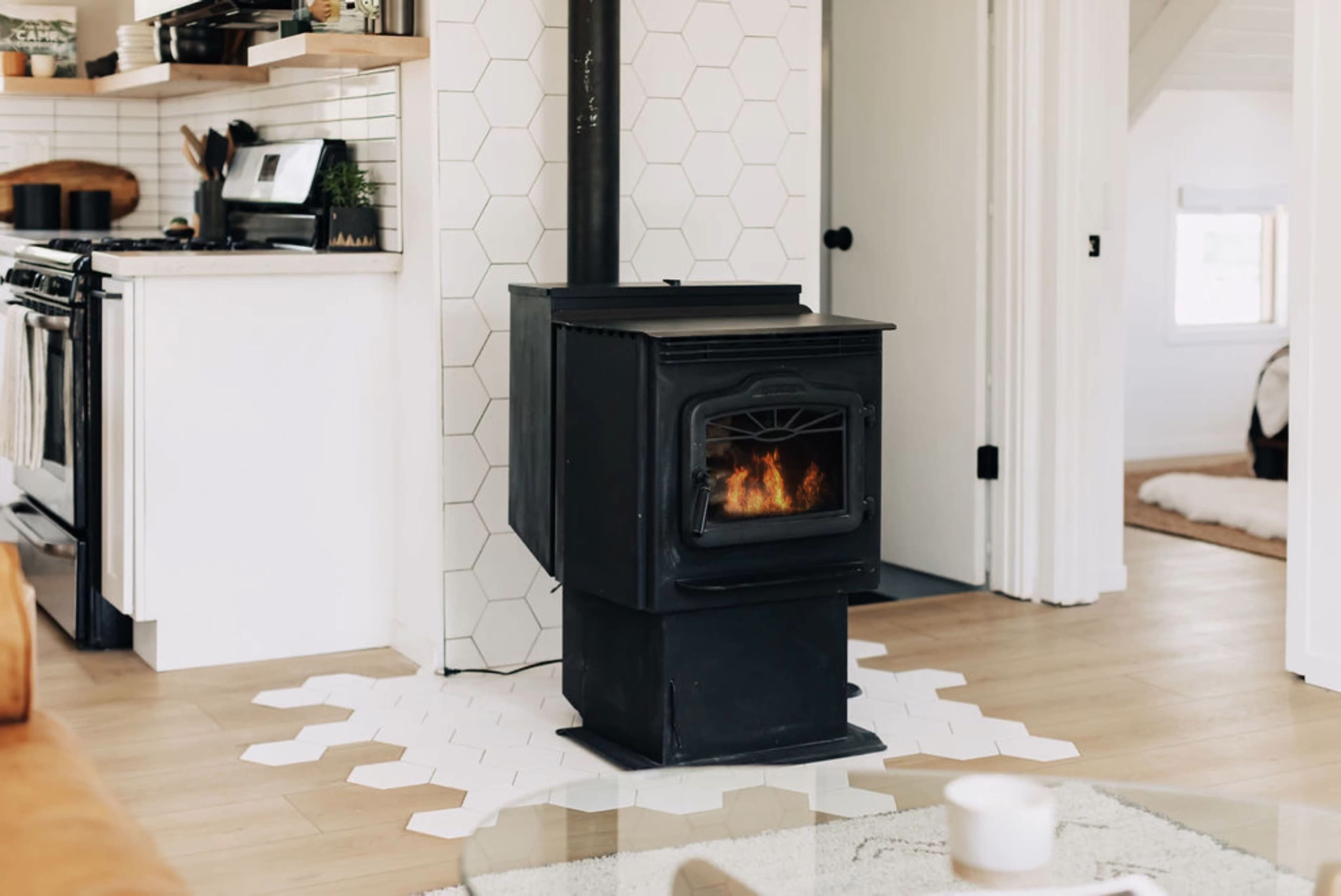 A black wood stove with a fire burning inside is centrally positioned on a white hexagonal tile base in a modern living room.