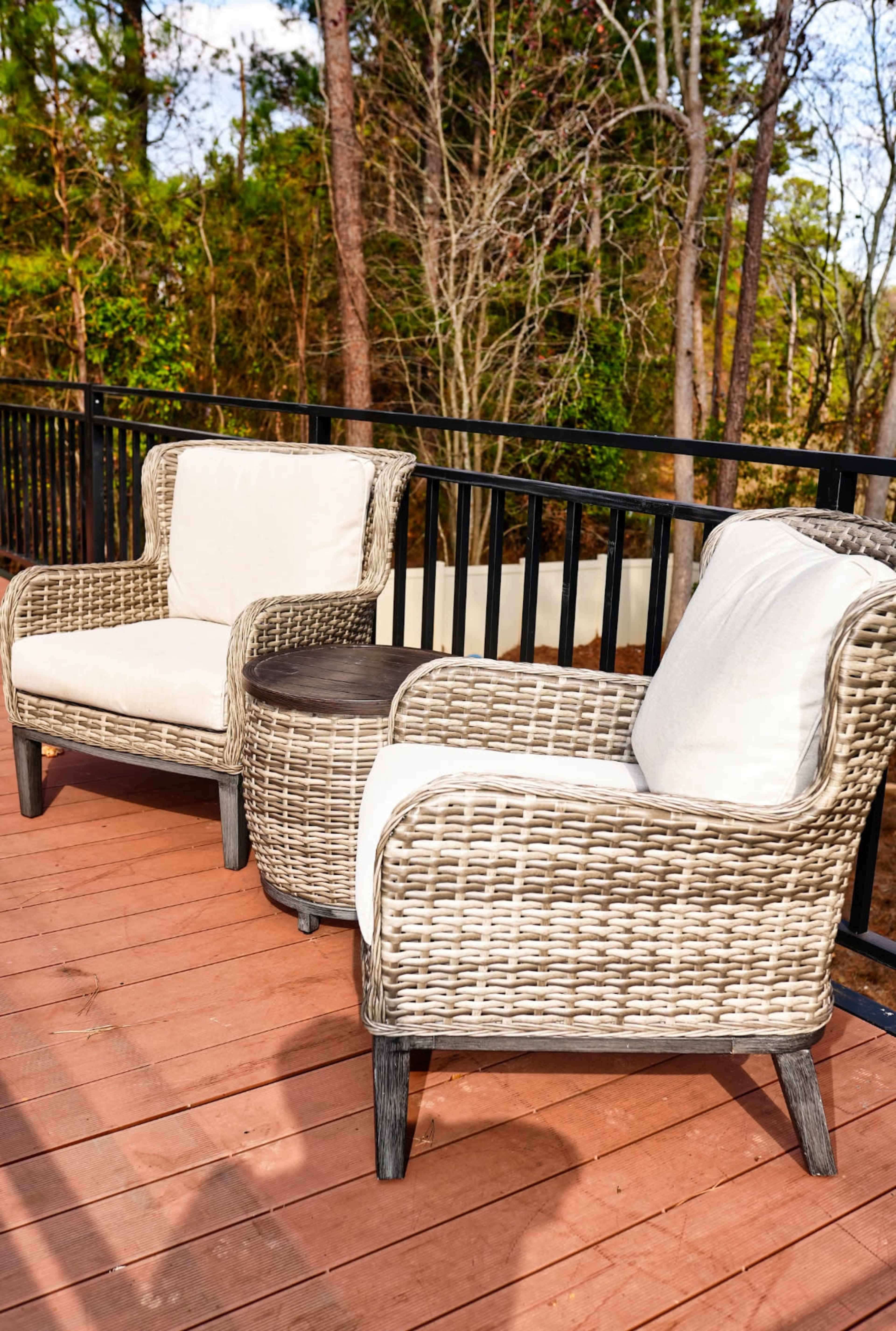 Two wicker chairs with white cushions and a small round table are positioned on a wooden deck surrounded by trees.