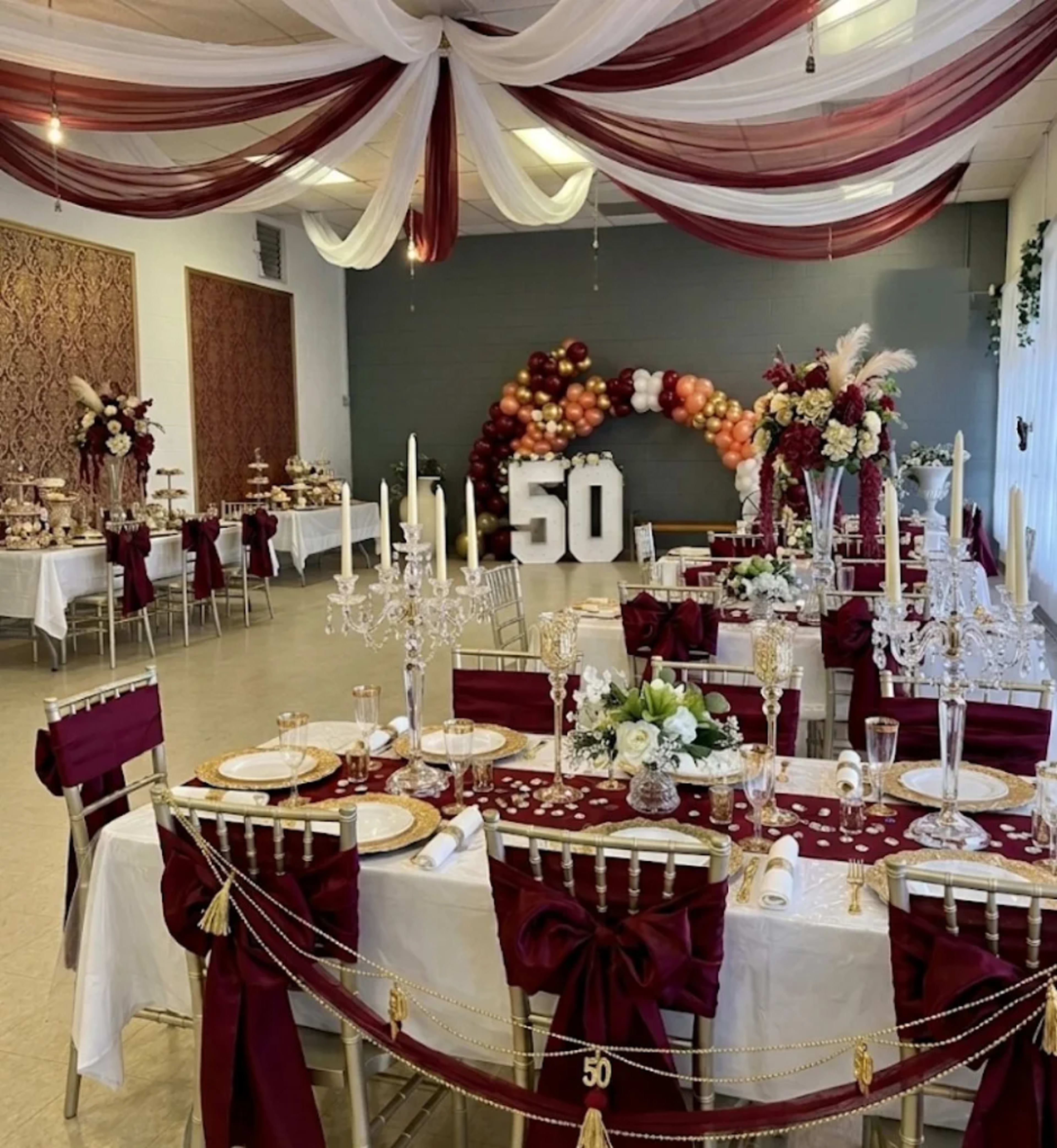 The image shows a elegantly decorated banquet hall for a 50th celebration, featuring tables with white tablecloths adorned with burgundy accents, floral centerpieces, and a backdrop of balloons and decor.
