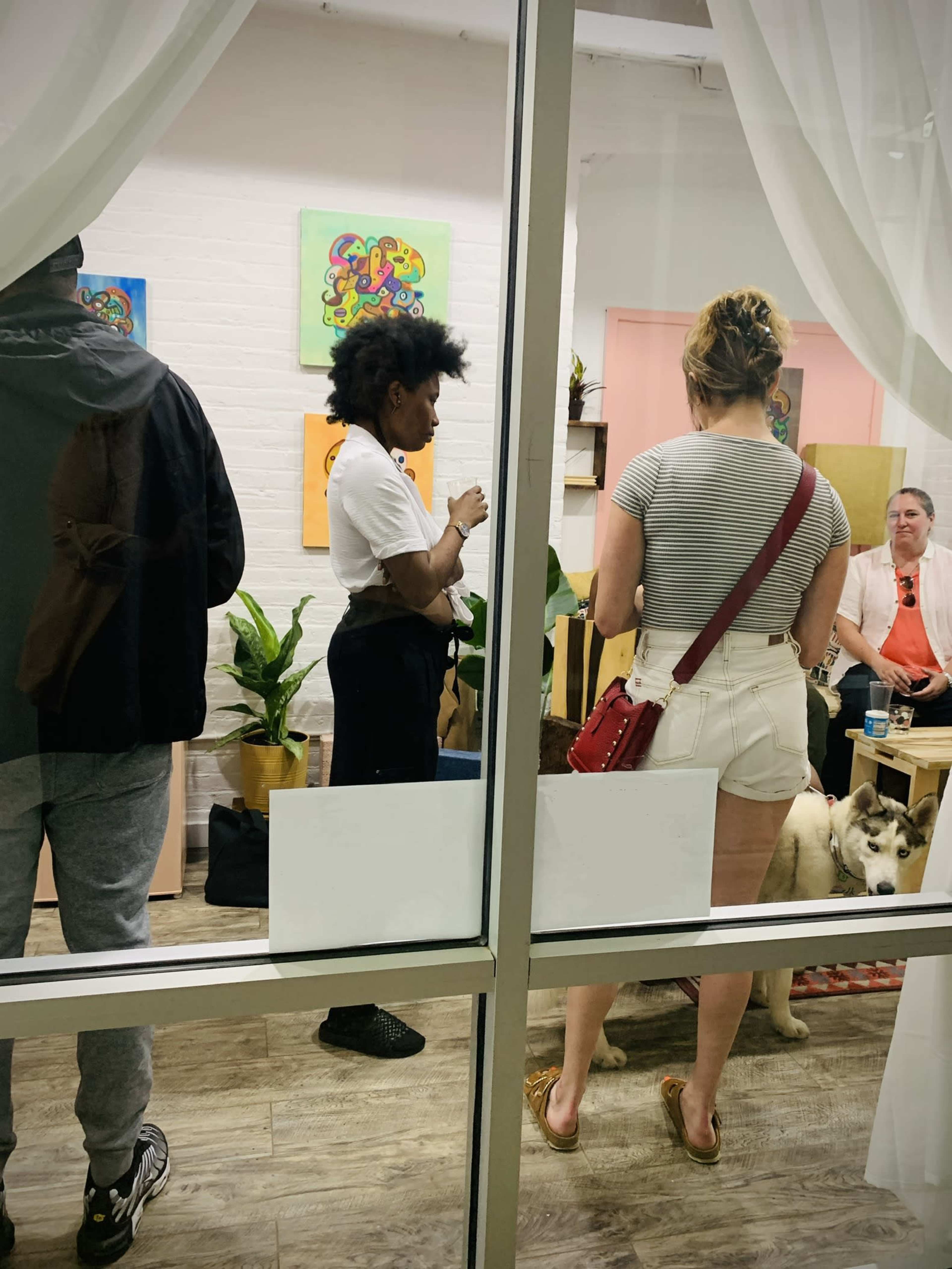 A gathering of people inside a room with plants, paintings on the walls, and a dog lying on the floor can be seen through a glass window.