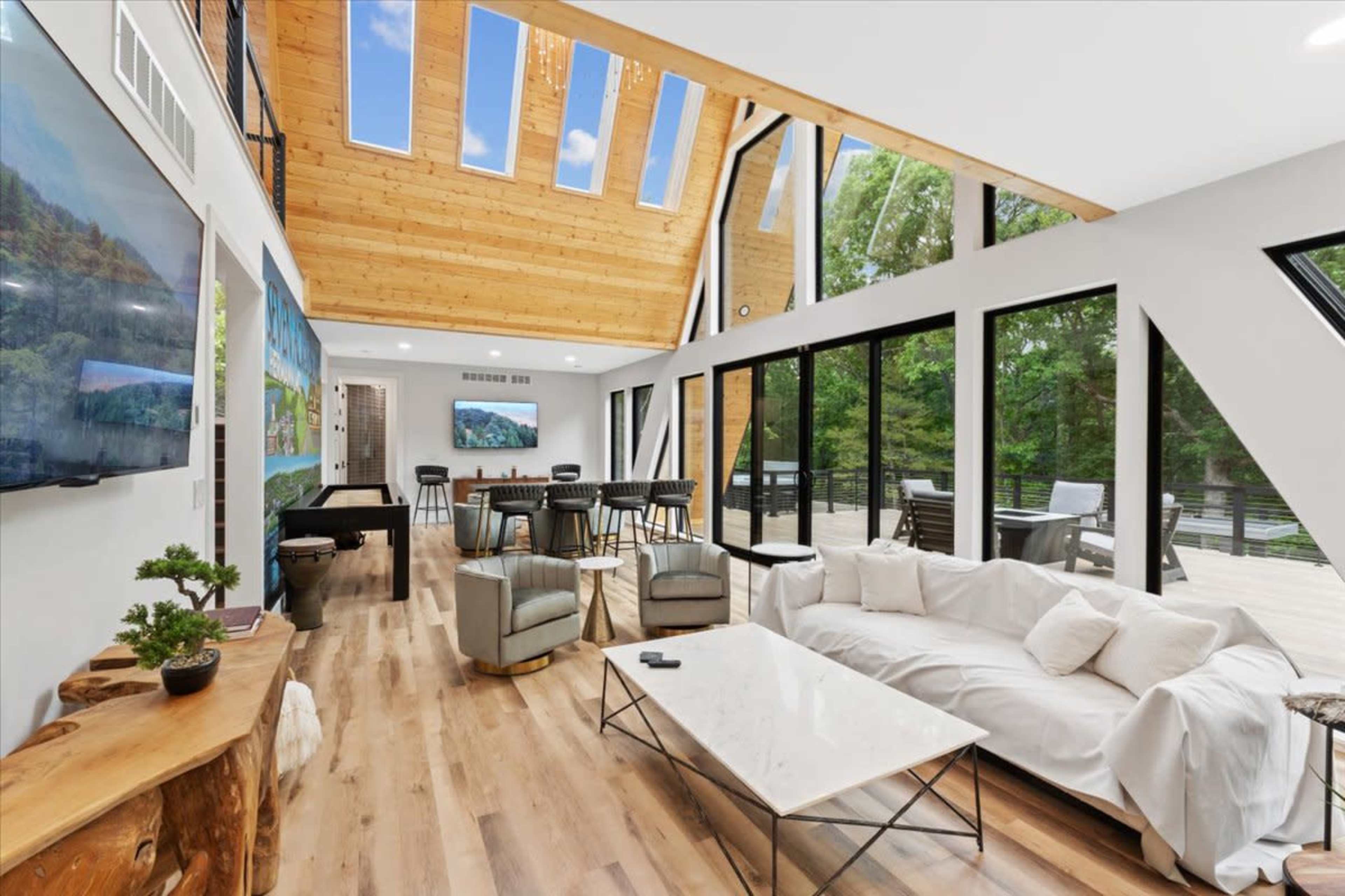 The image shows a modern living room with large windows, a white couch, a marble coffee table, and a wooden ceiling, overlooking a deck and natural surroundings.