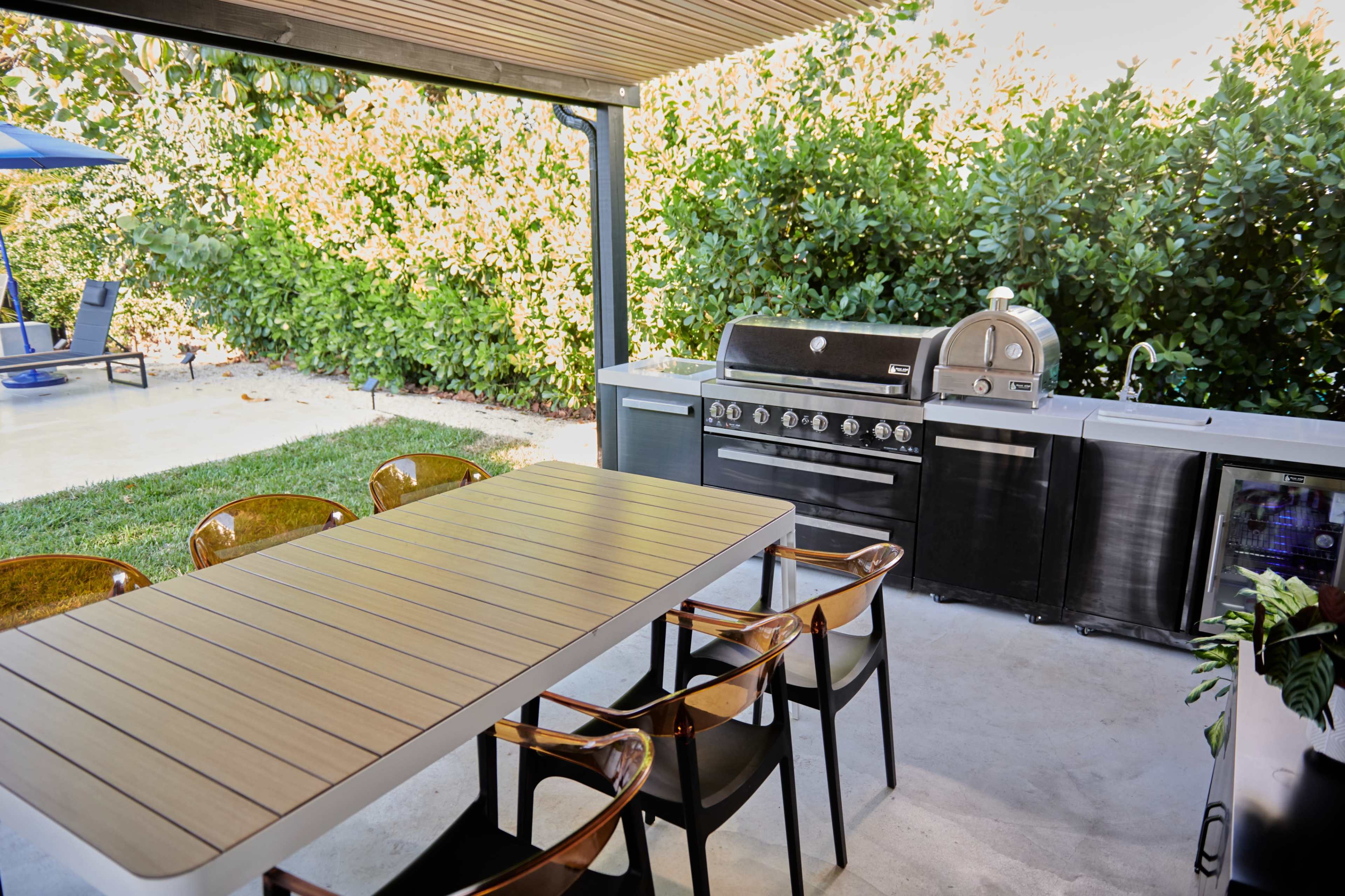 An outdoor kitchen area featuring a grill, a pizza oven, a sink, and a dining table with chairs under a covered structure, surrounded by greenery.