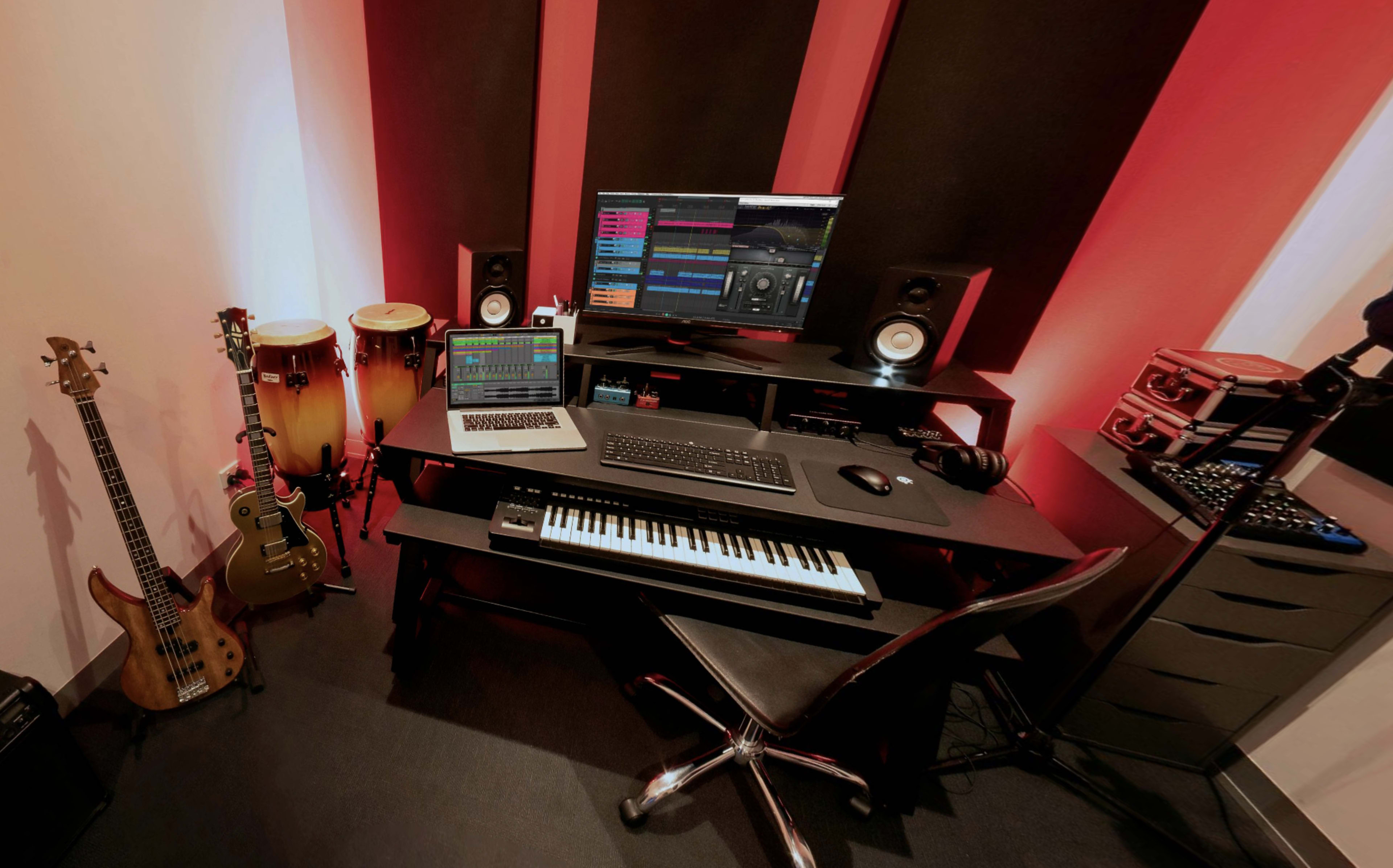 A compact music studio features a computer workstation with software displayed, musical instruments including guitars and congas, and sound equipment arranged around a black desk.