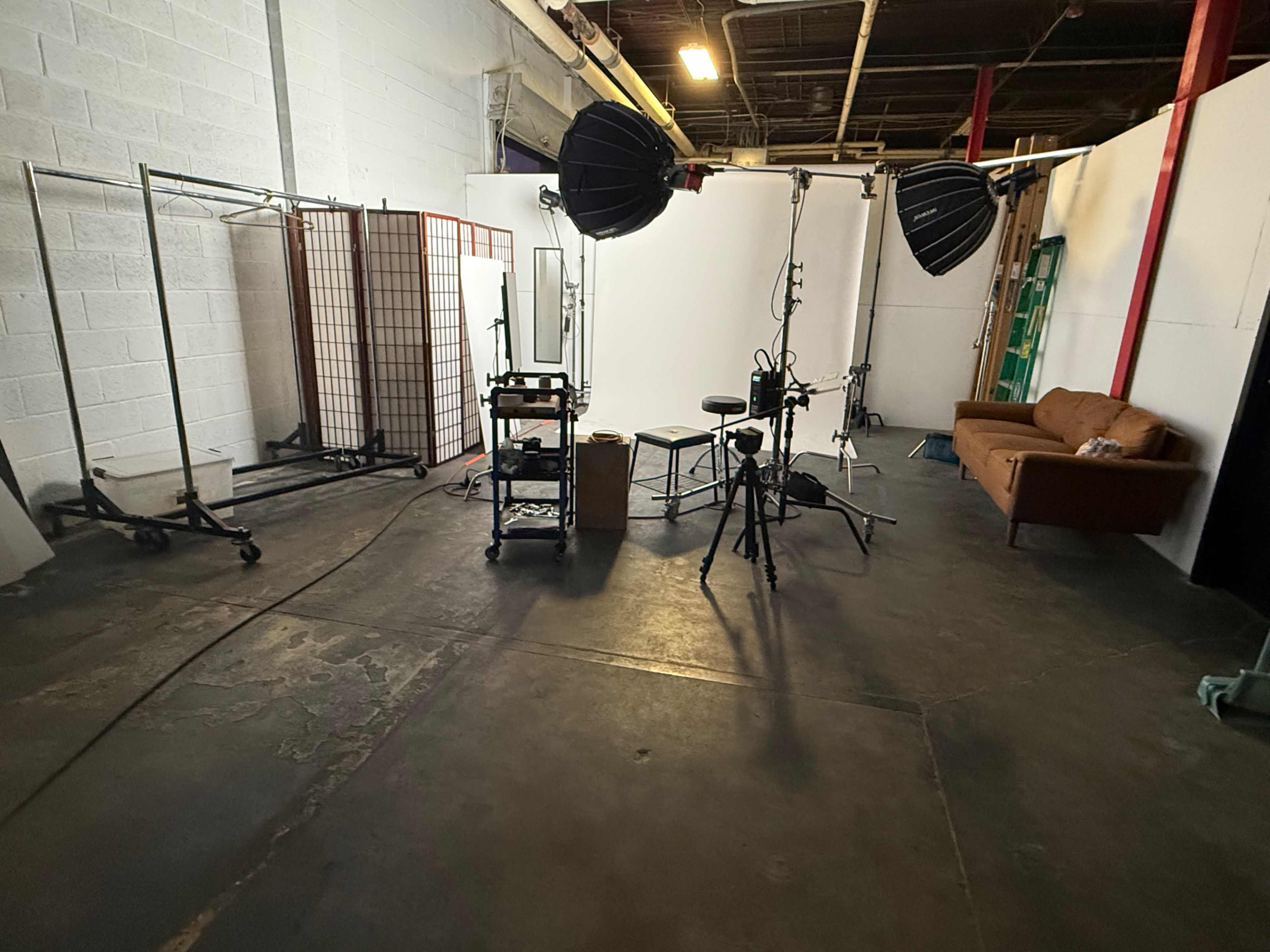The image shows a photography studio with various equipment including lights, a tripod, and a sofa, set against a backdrop of white walls.