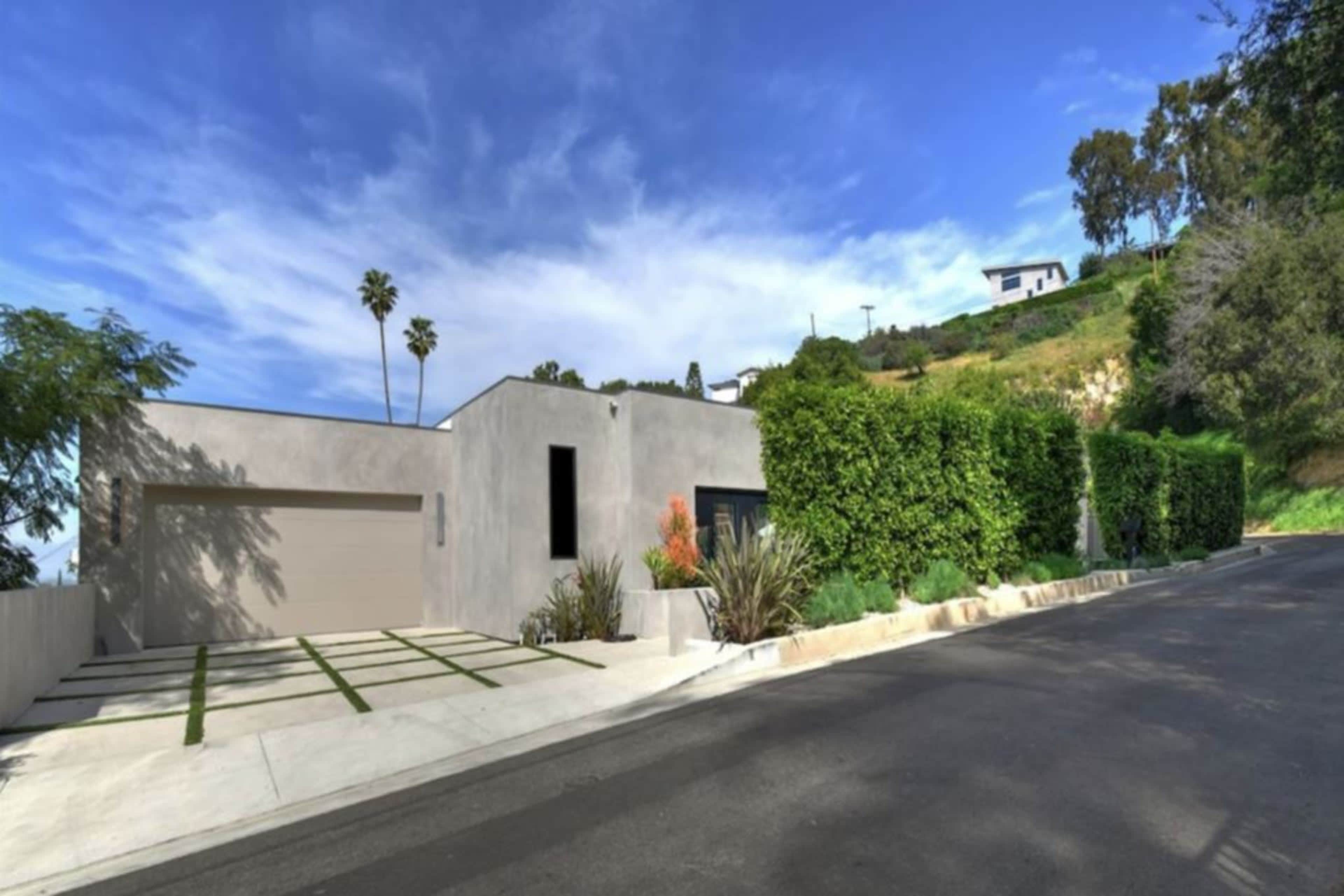 The image shows a modern house with a landscaped front yard and a sloped driveway, surrounded by greenery on a hillside.