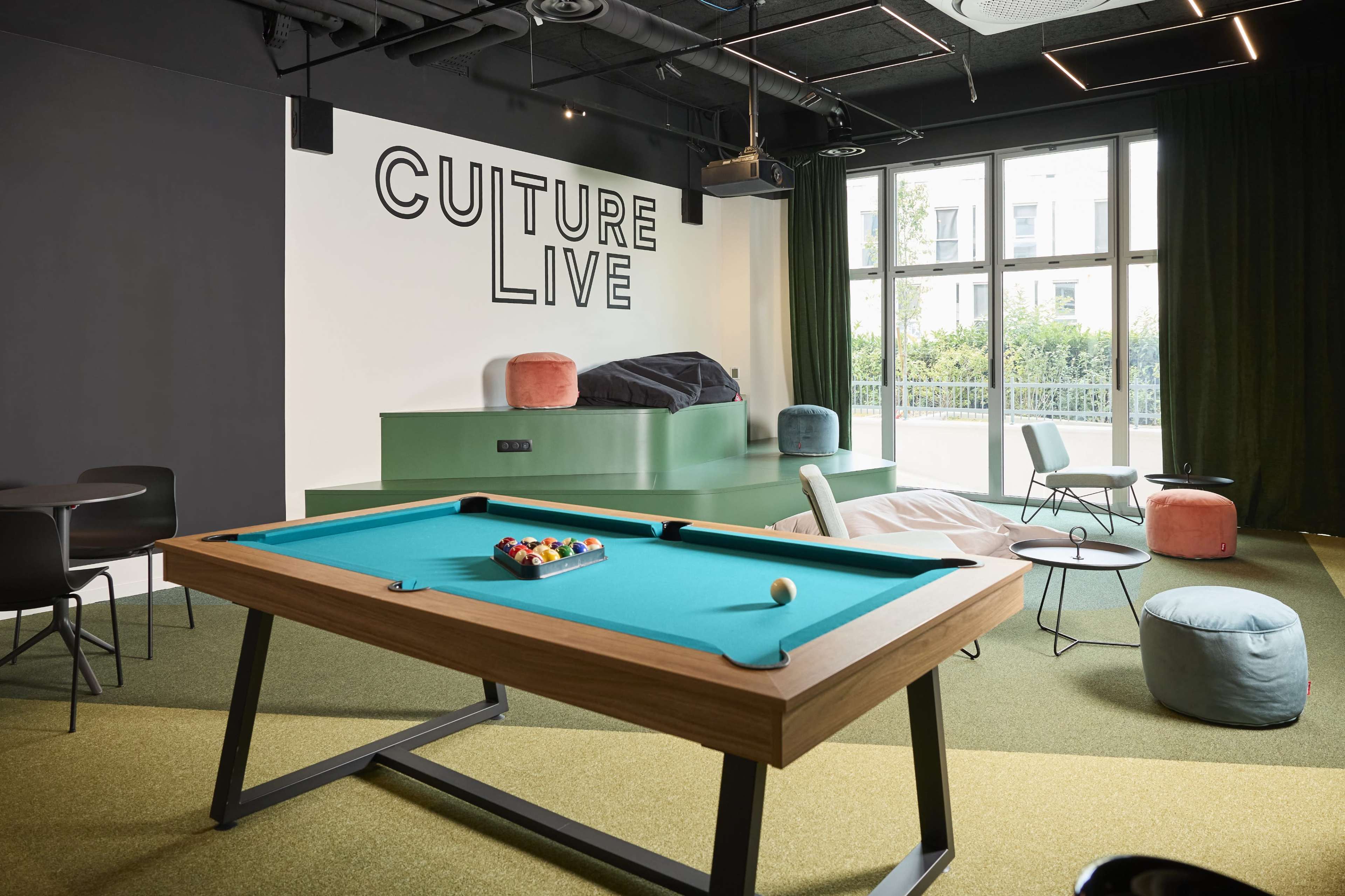 The image shows a modern recreational area featuring a green pool table in the foreground and a large wall with the text "CULTURE LIVE" in bold letters, along with seating arrangements and colorful bean bags in the background.