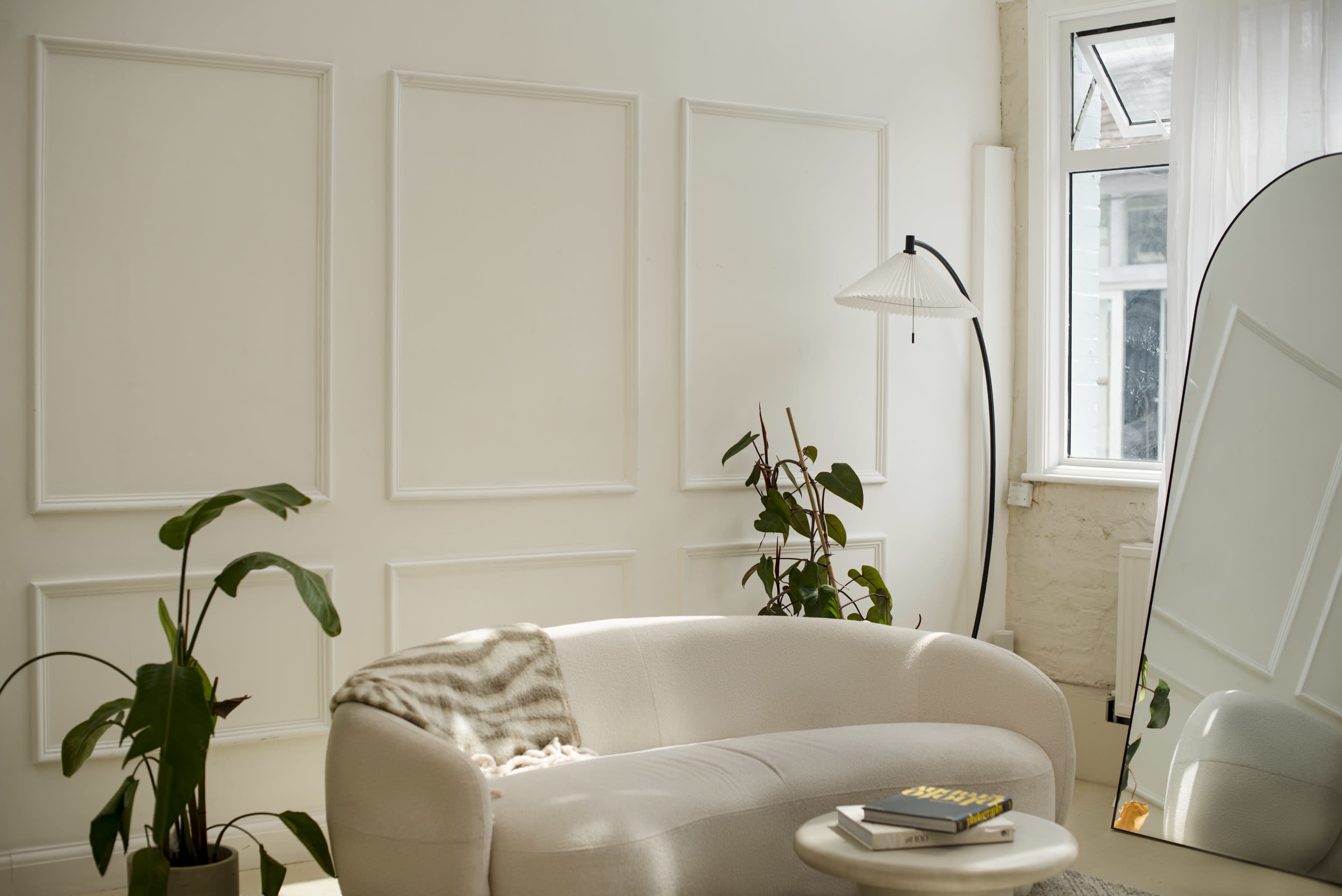 The image shows a bright, modern living room featuring a curved beige sofa, a floor lamp, several indoor plants, and a large mirror reflecting natural light from a window.
