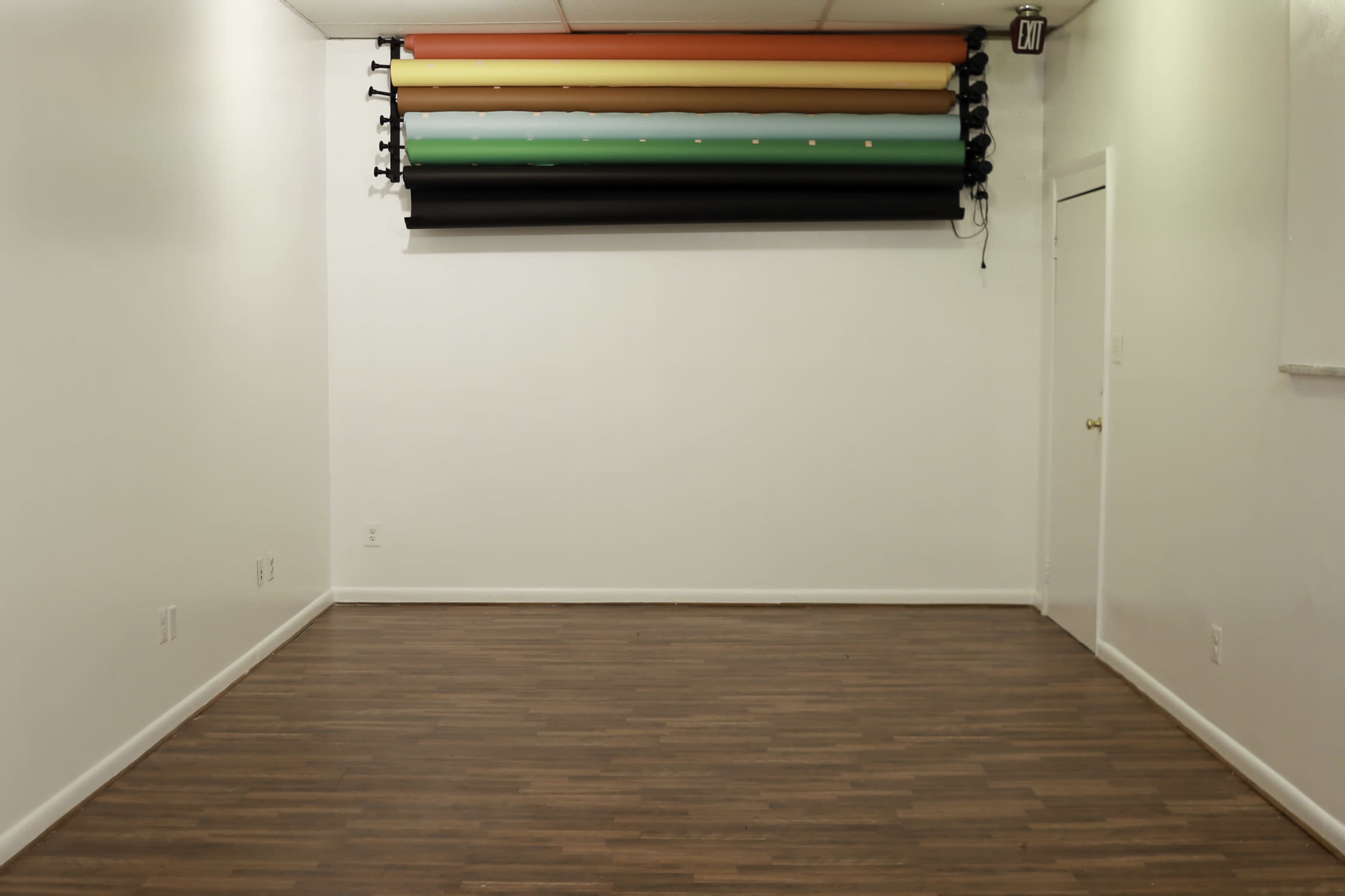 The image shows a minimalistic studio space featuring a wooden floor and a wall-mounted backdrop with horizontal colored rolls above it.