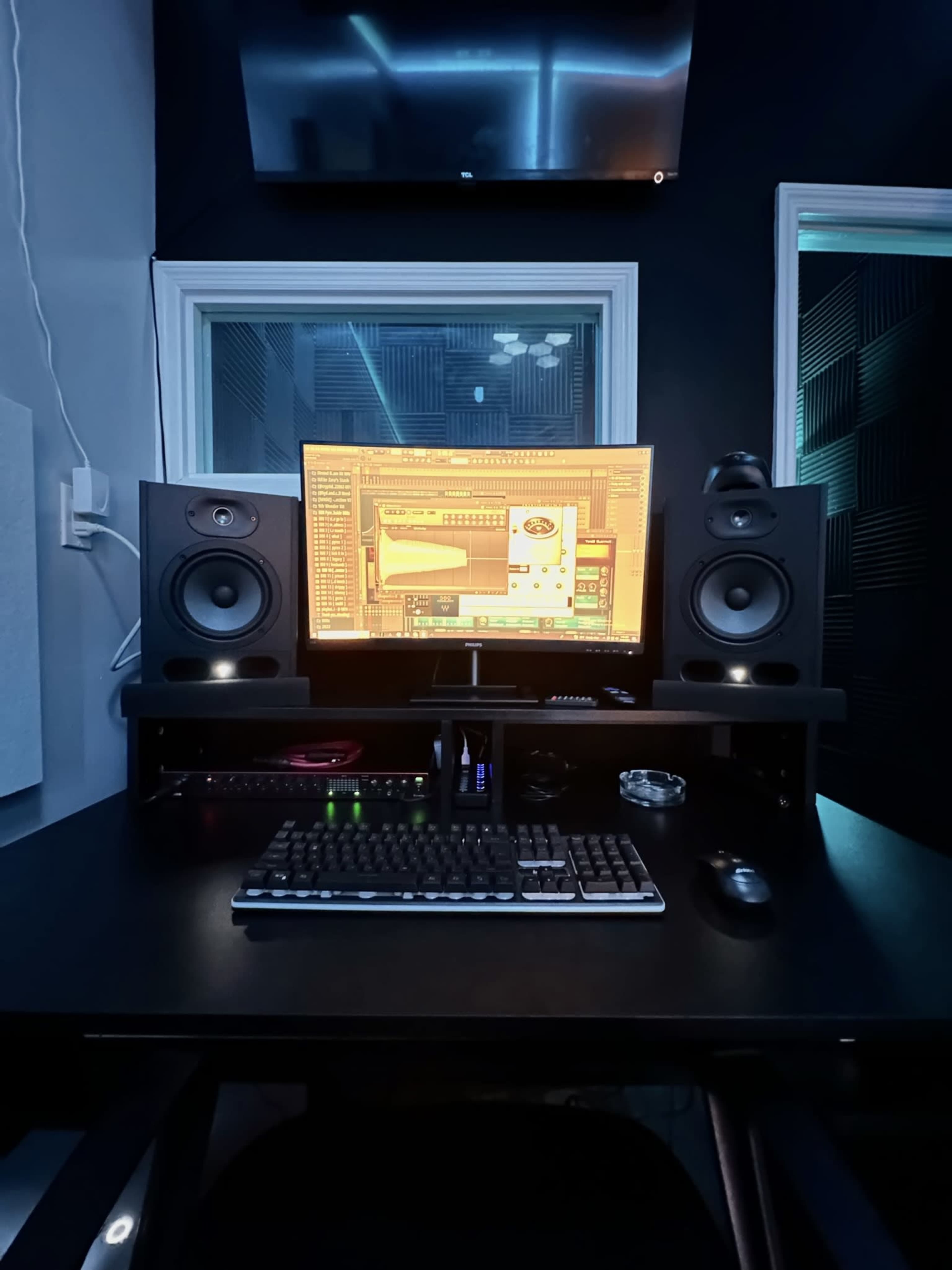 A music production workstation features a large monitor, studio speakers, a keyboard, and a mouse set against a dark wall with soundproofing panels.