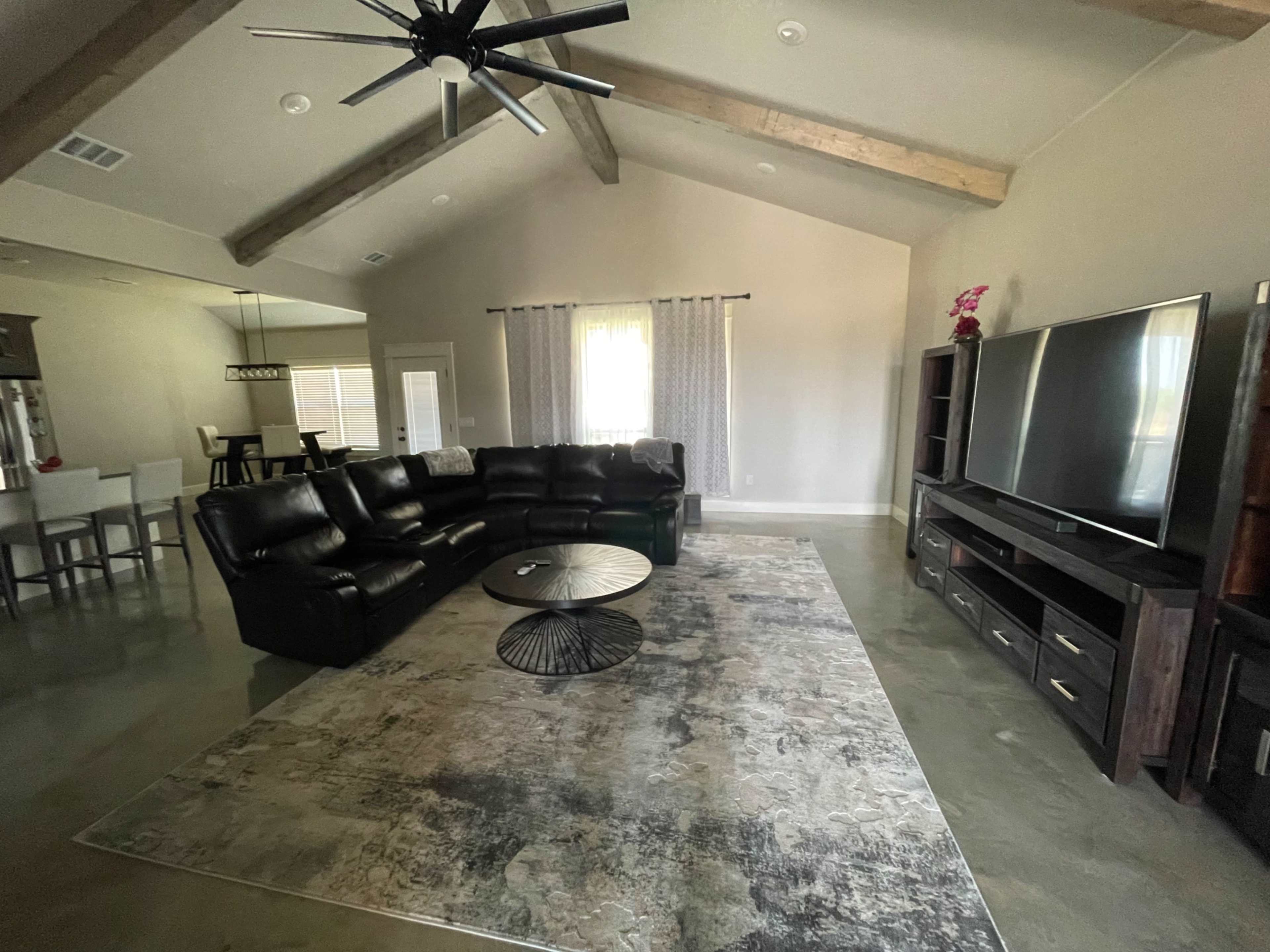 A spacious living room with a black sectional sofa, a round coffee table on a large area rug, and a wall-mounted television.