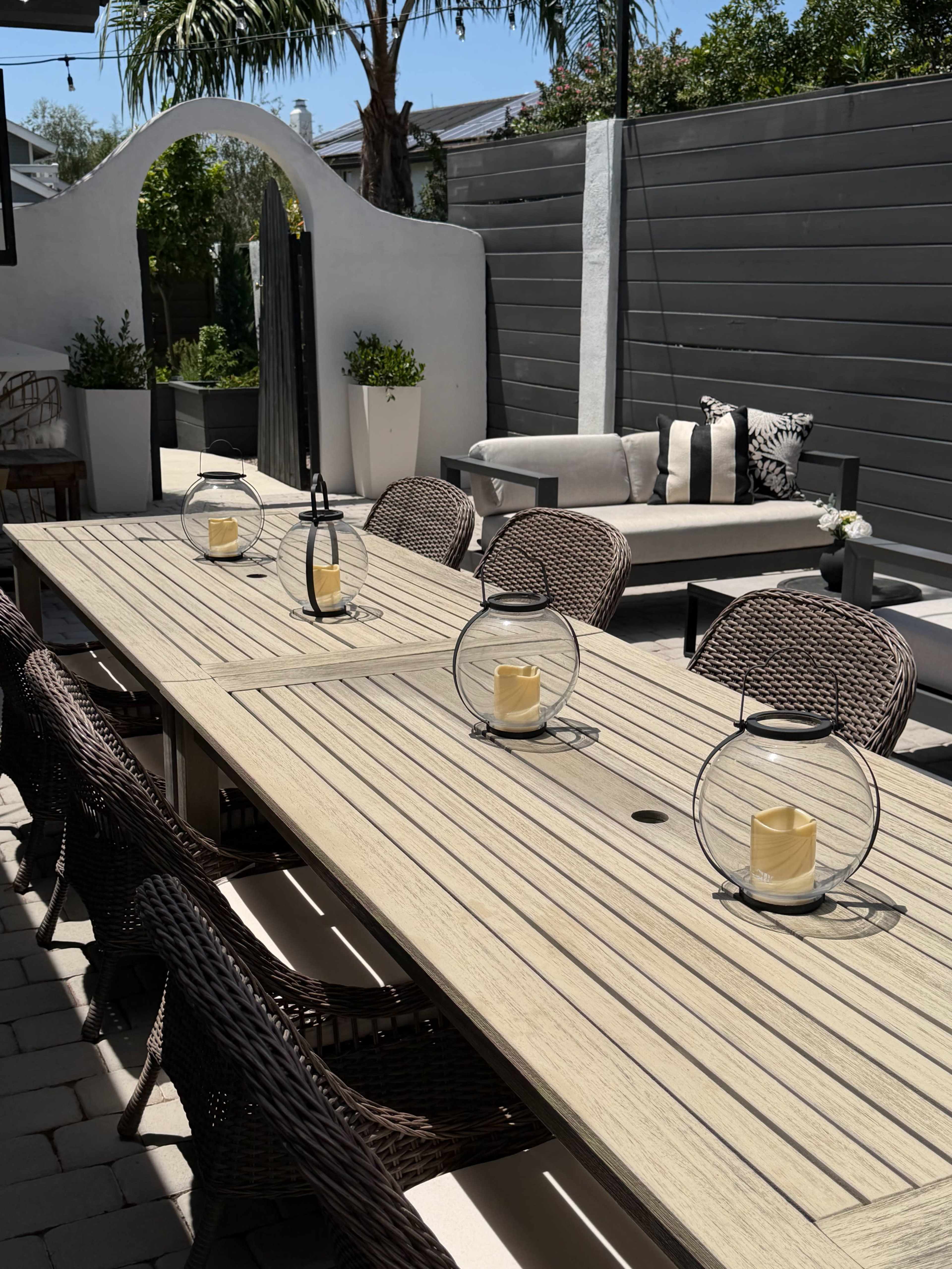 A long wooden dining table with several wicker chairs is set outdoors, featuring lanterns with candles on top, surrounded by a patio area with a white arch and potted plants.