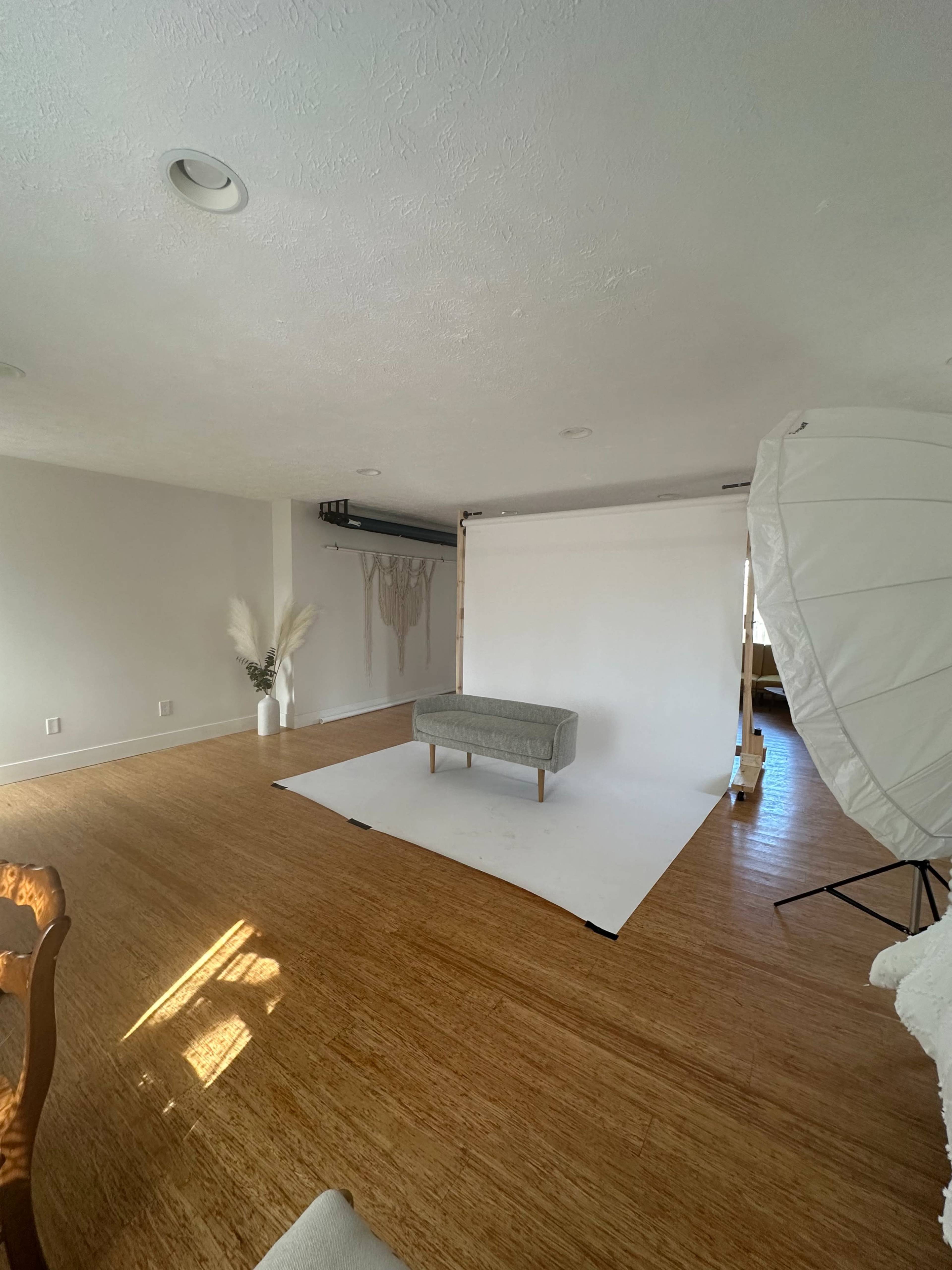 A spacious room with a white backdrop, a gray bench in front, and photography equipment set up on the side.