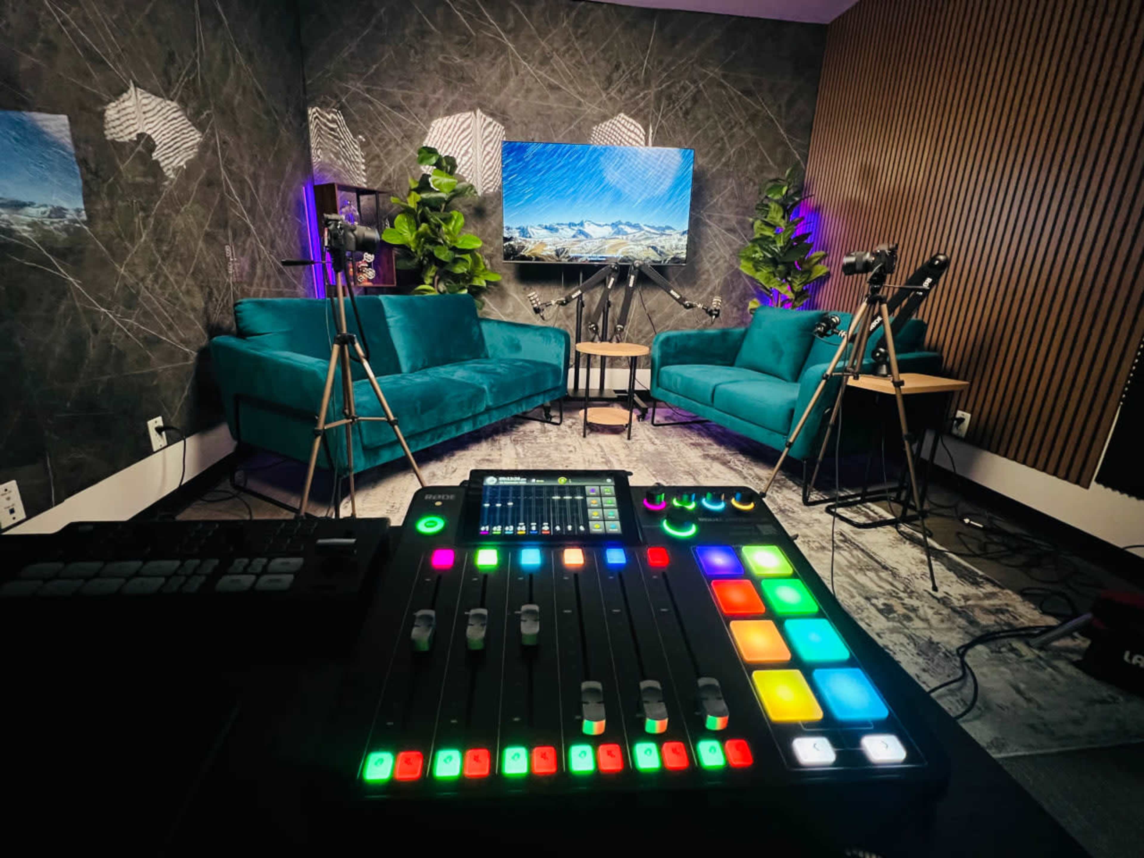 The image shows a modern video production studio featuring teal couches, tripods with cameras, and a colorful audio mixing console prominently in the foreground.