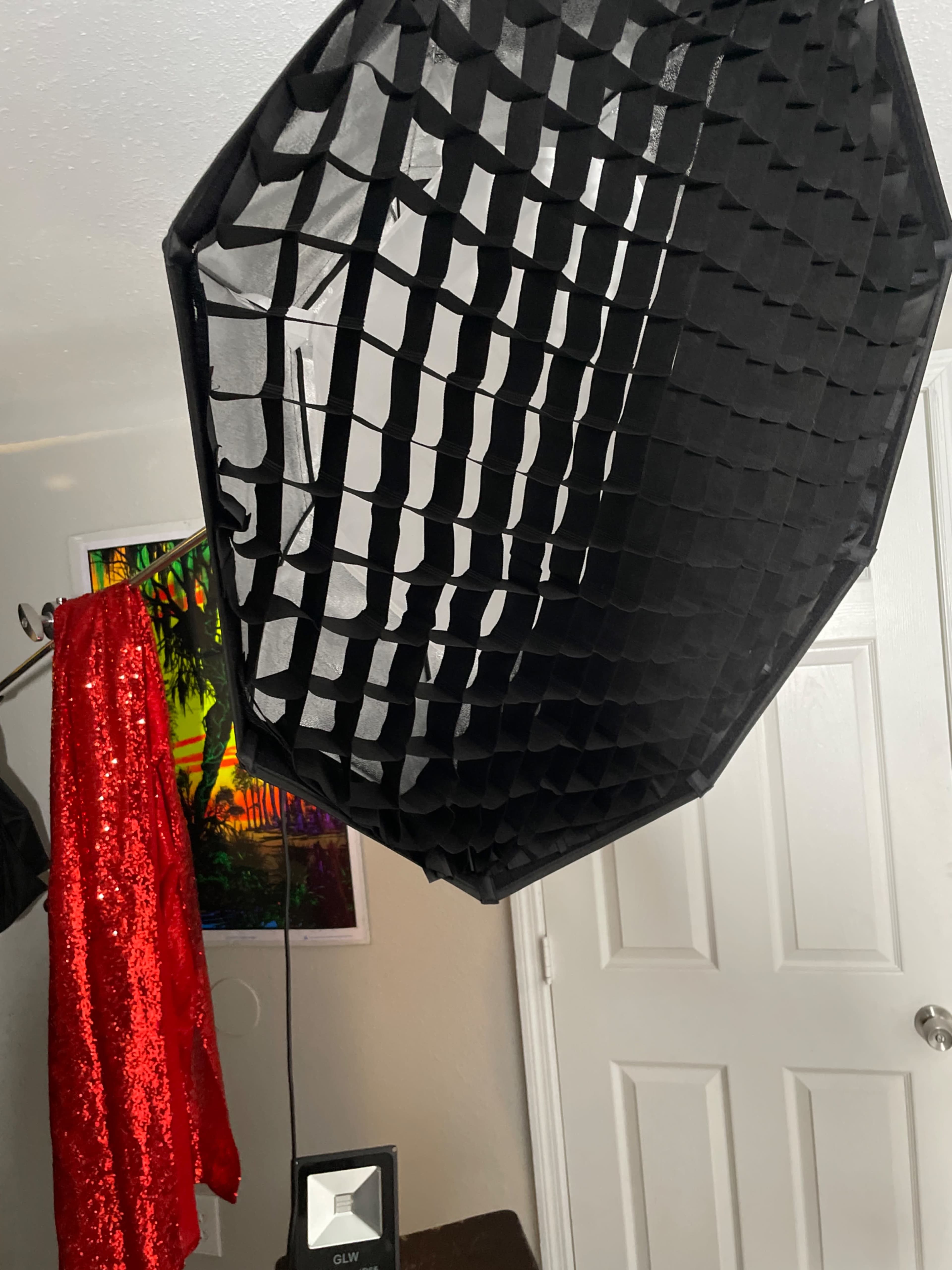 A black softbox with a grid set up in a room, alongside a shiny red garment hanging on a rack and a door in the background.