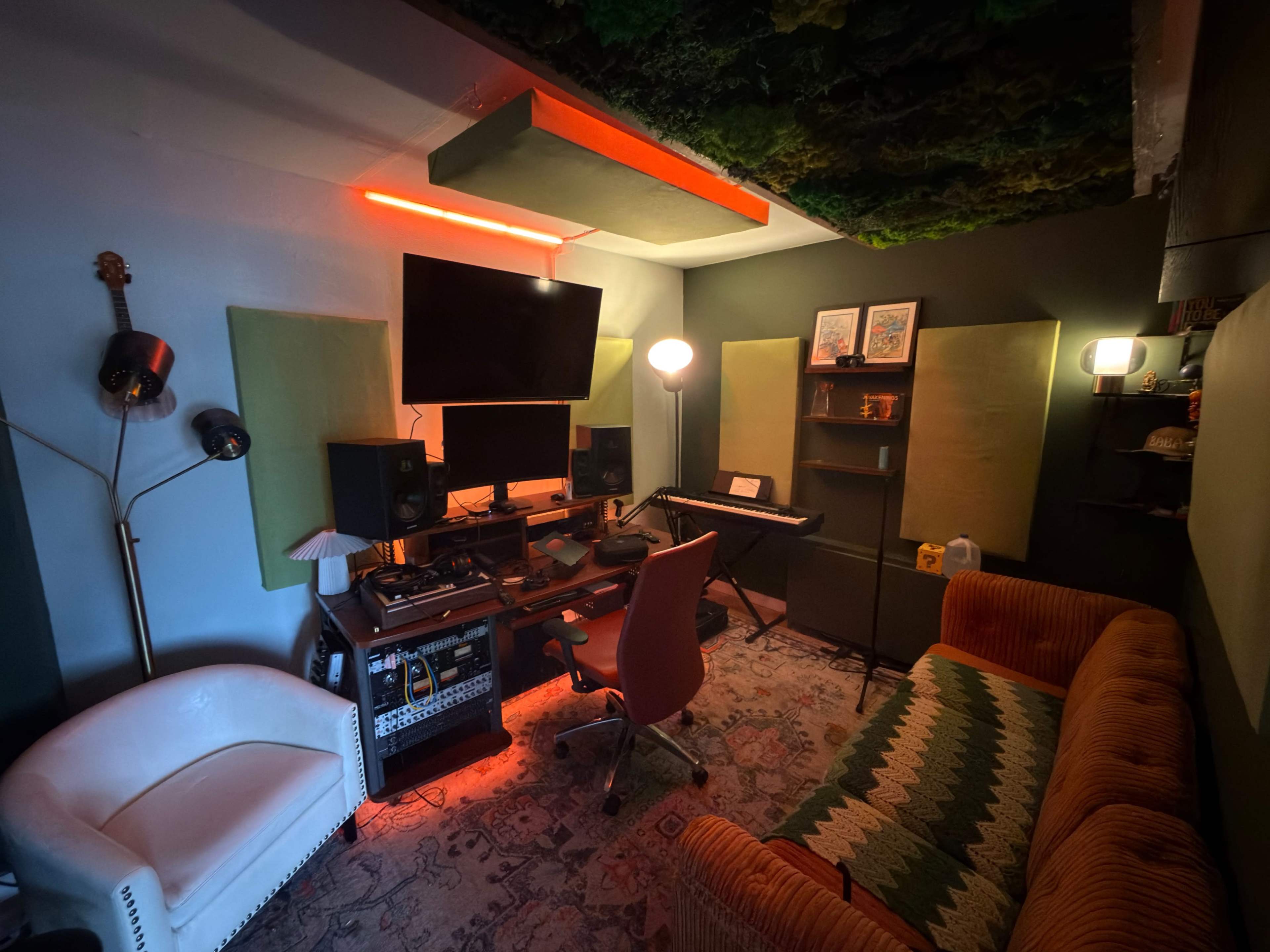 The image shows a small music studio featuring a mixing desk, a keyboard, acoustic panels, and comfortable seating.
