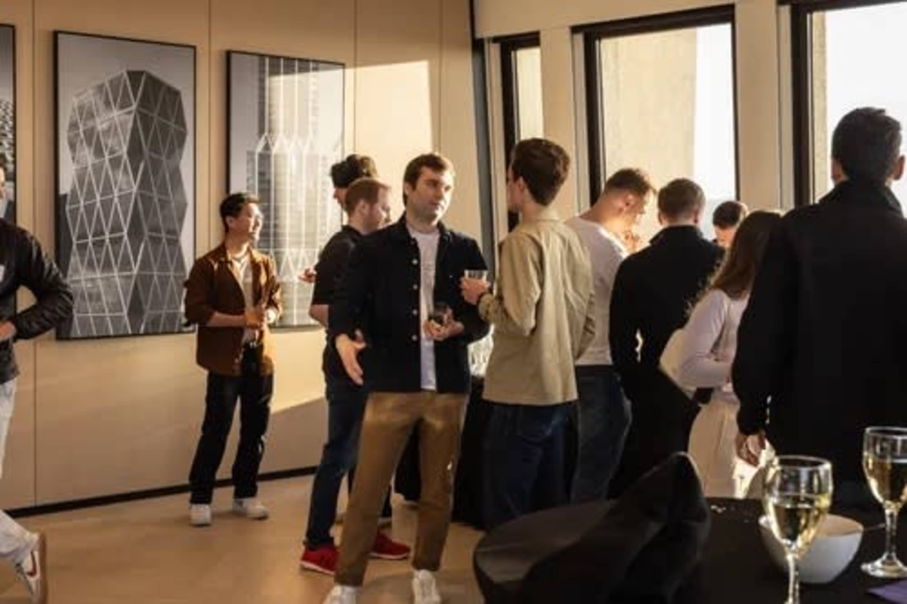 A group of people socialize in a modern room with large windows and geometric artwork on the walls.