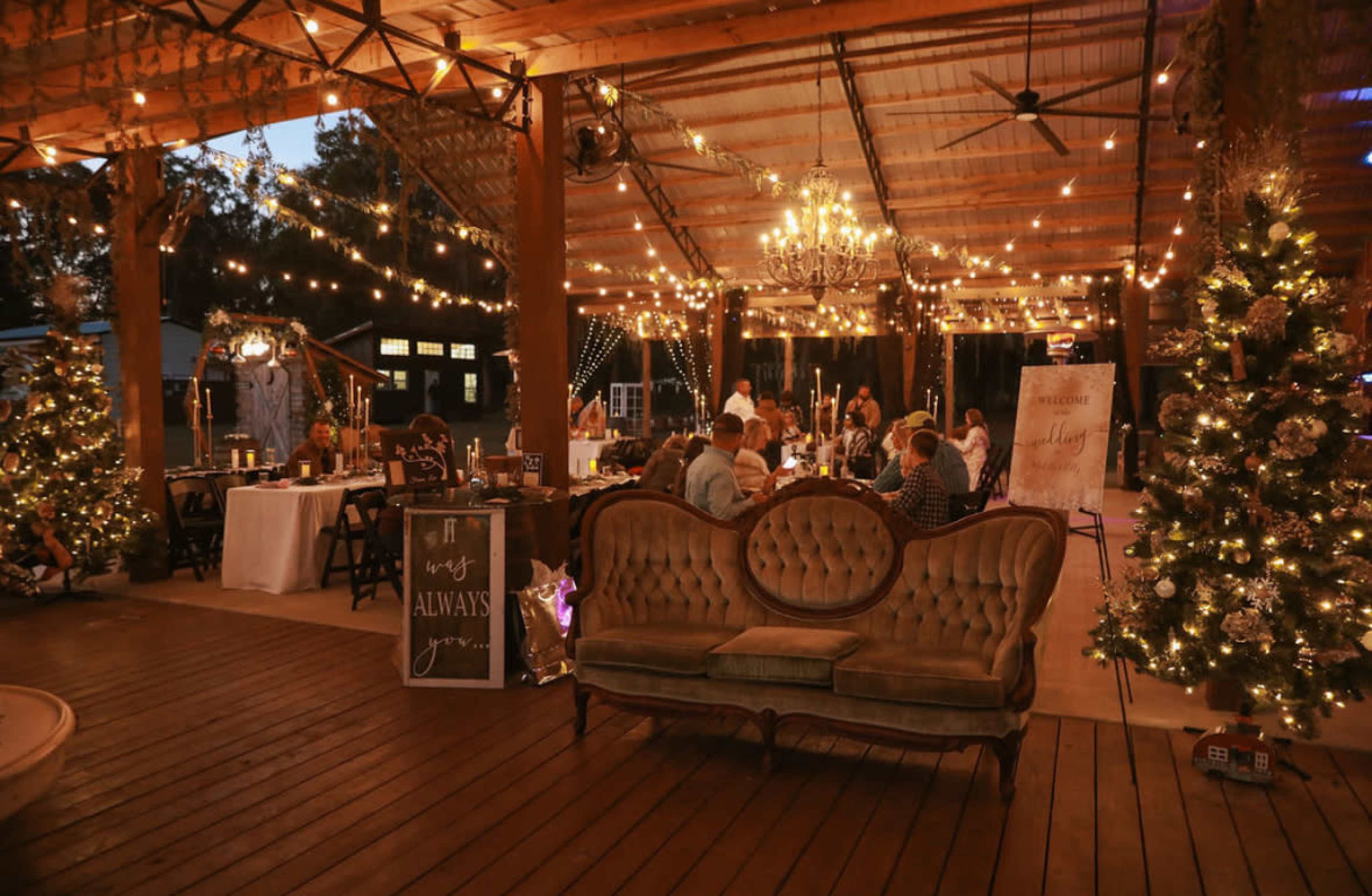 The image shows a beautifully decorated open space with string lights, Christmas trees, and a vintage couch, set for an event.