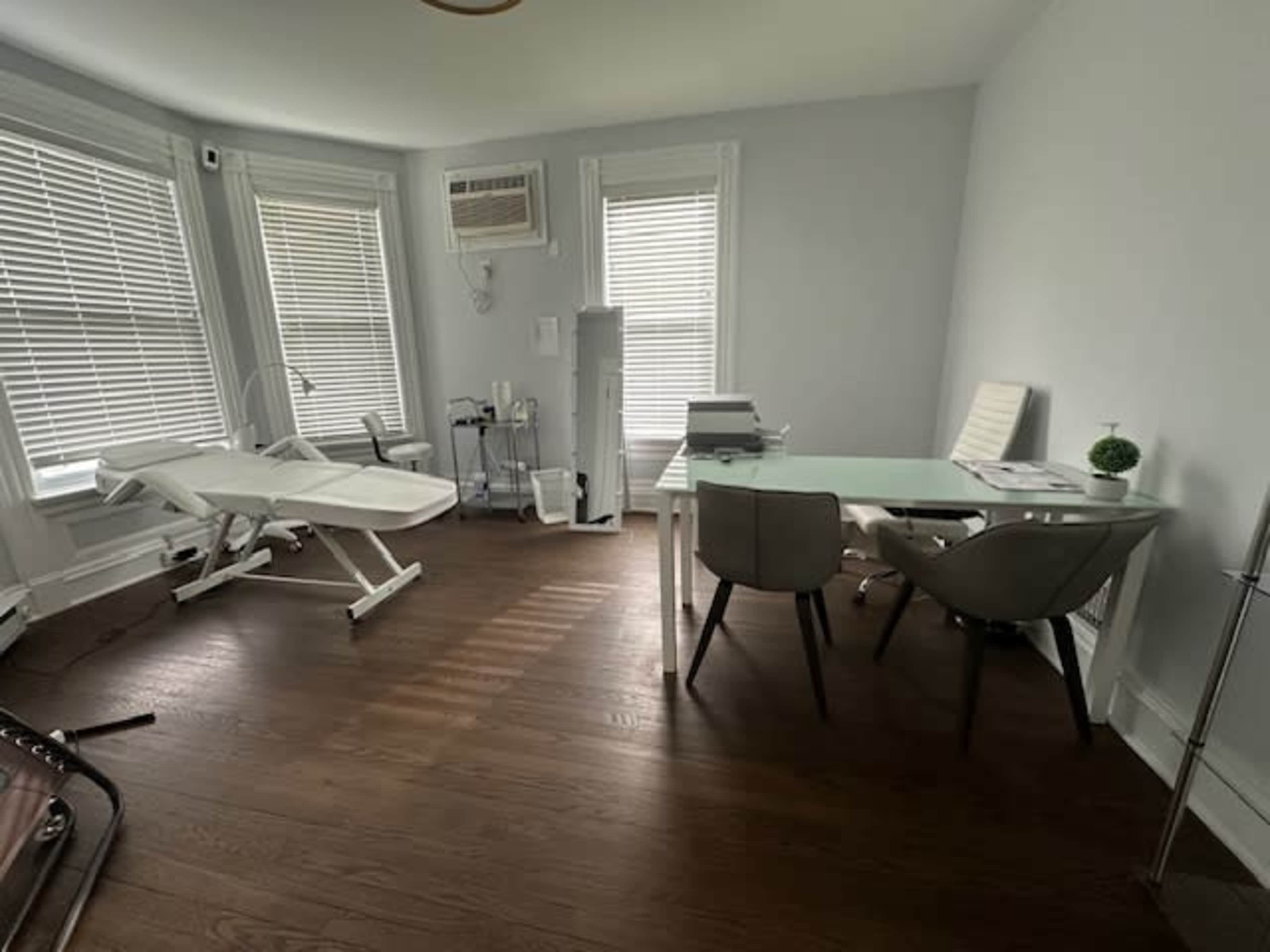 A well-lit office space with a treatment bed, a desk with chairs, and a small plant on the table, featuring wooden flooring and large windows.