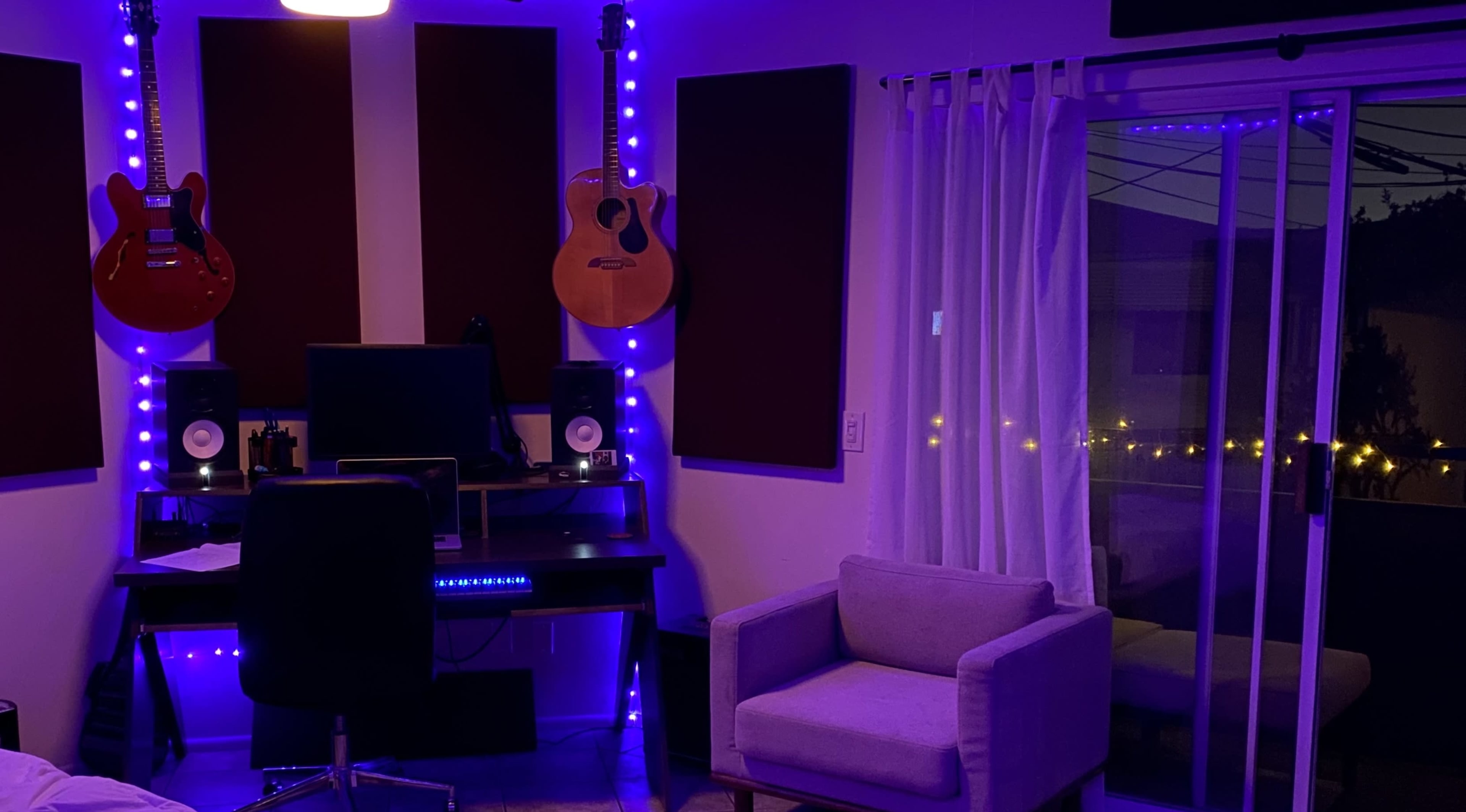 The image shows a music studio with an acoustic guitar hanging on the wall, a desk with studio monitors, a chair, and a large window with light curtains.