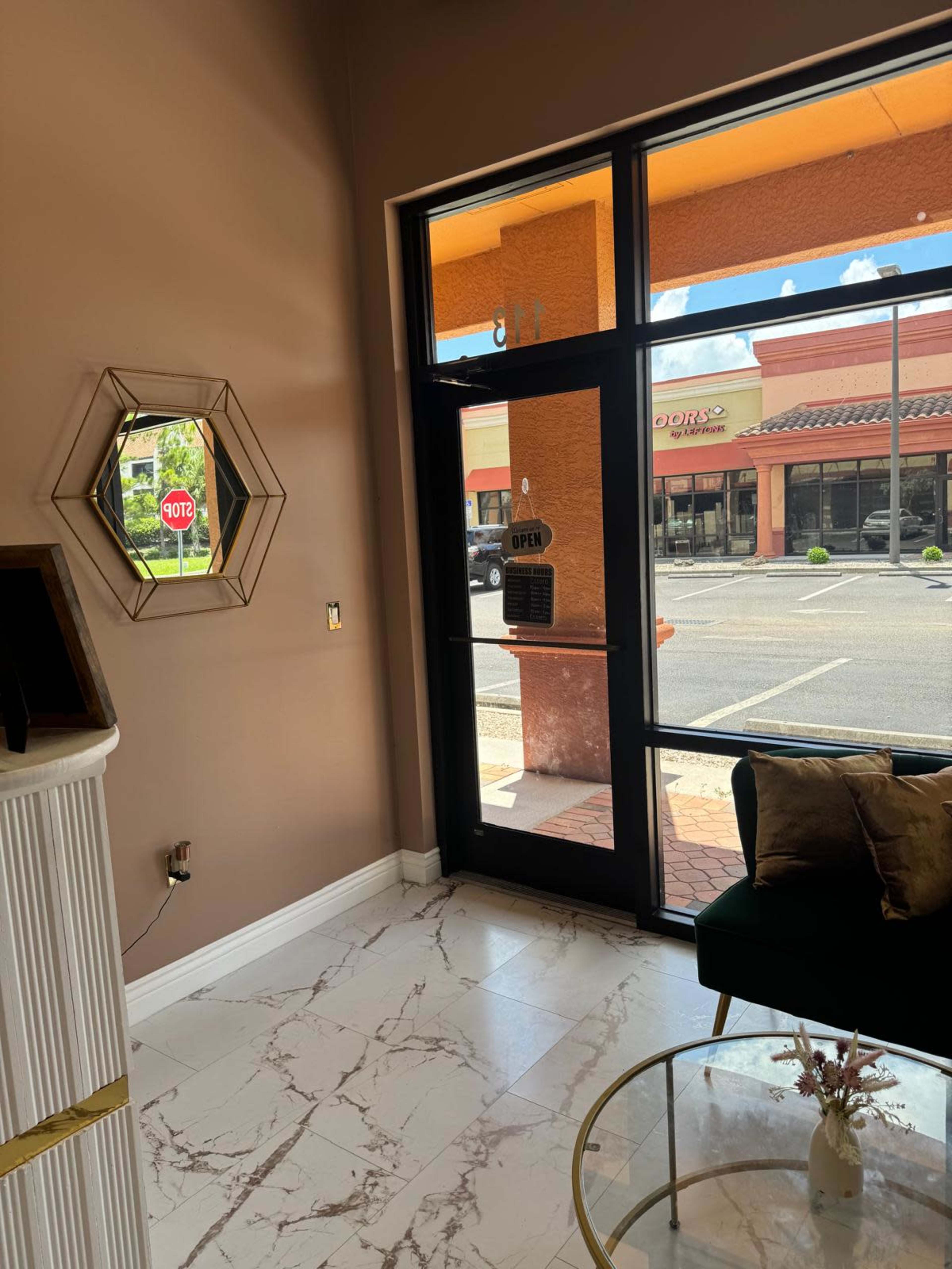 The image shows a well-lit interior of a shop featuring a decorative mirror, a green couch, and a glass coffee table, with a view of the parking lot through large windows.