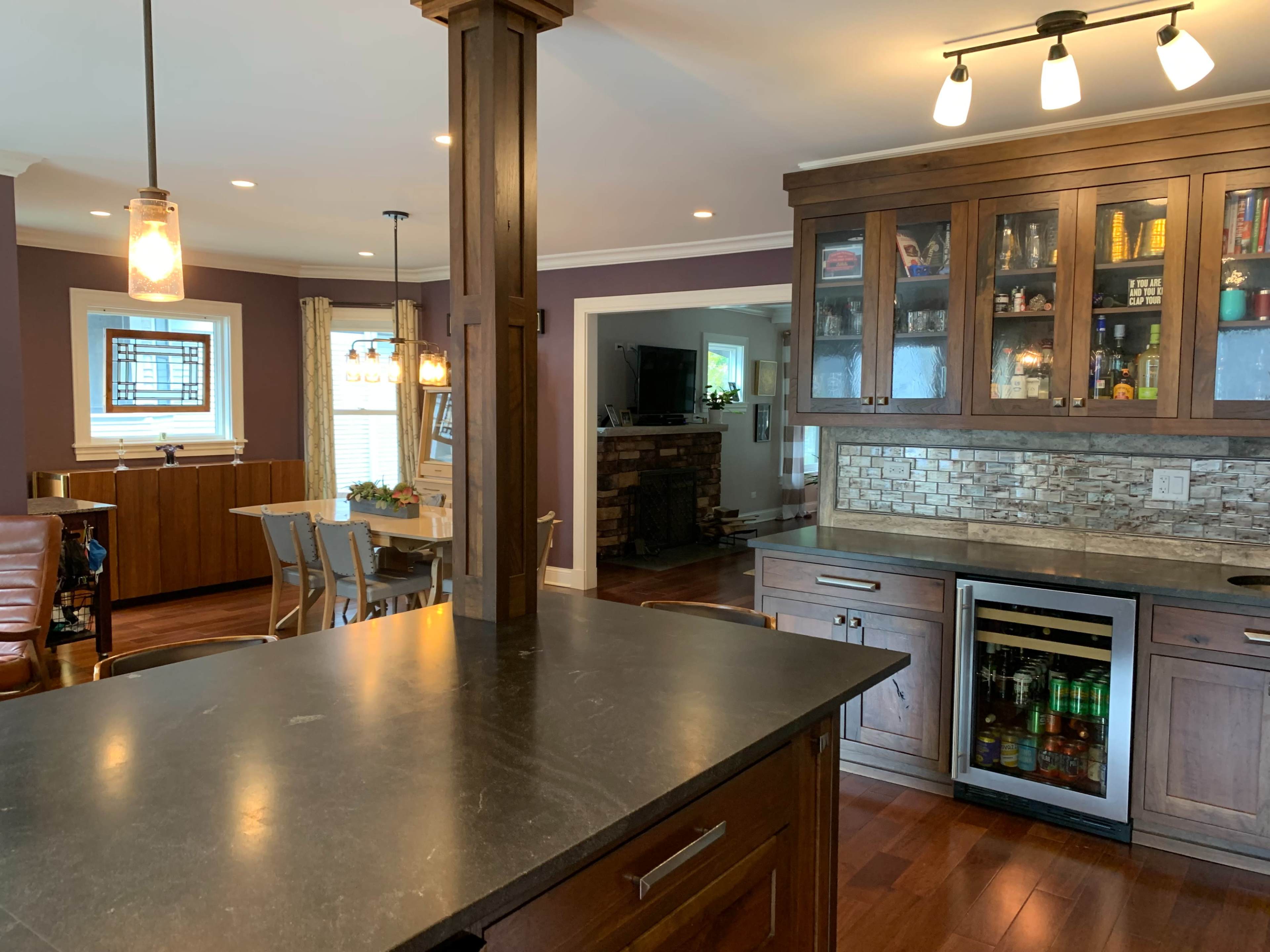 A modern kitchen with dark wooden cabinets, a black countertop, and a glass-fronted cabinet displaying various items, with a dining area visible in the background.