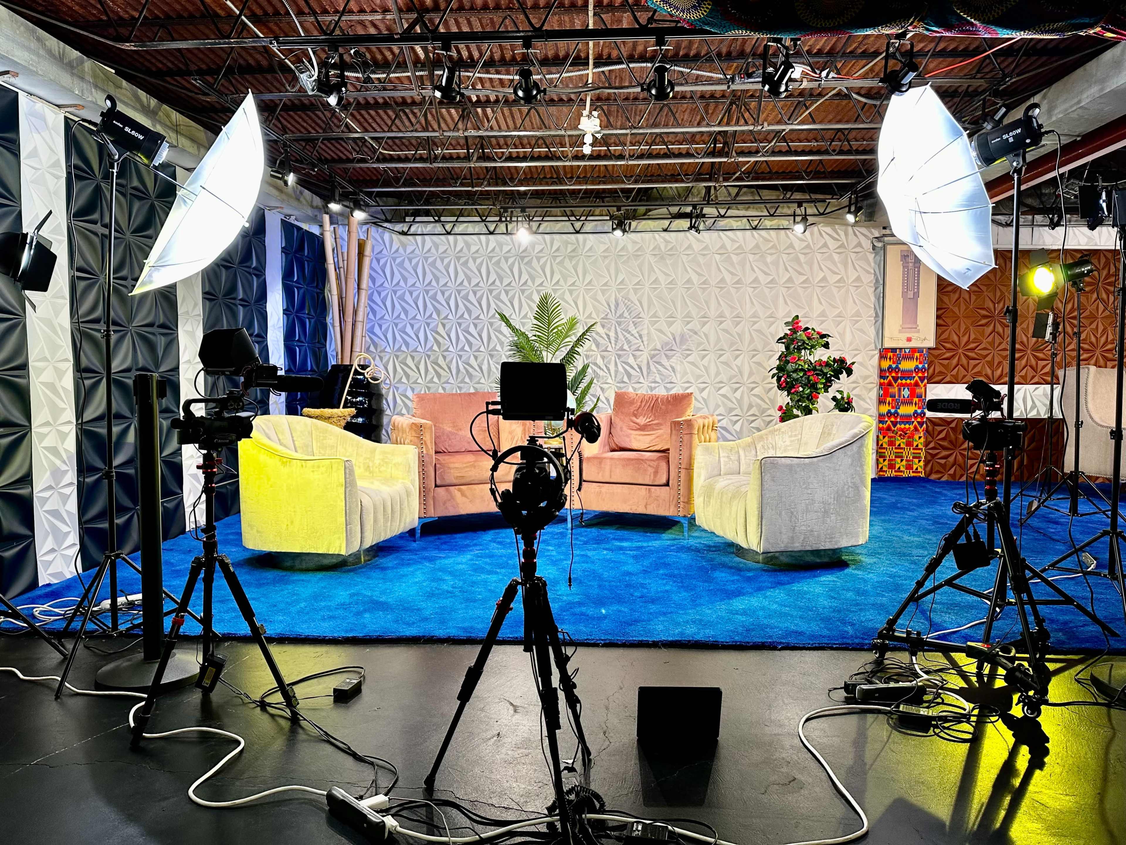 A television studio setup with two pastel-colored couches on a blue carpet, surrounded by camera equipment and lighting.