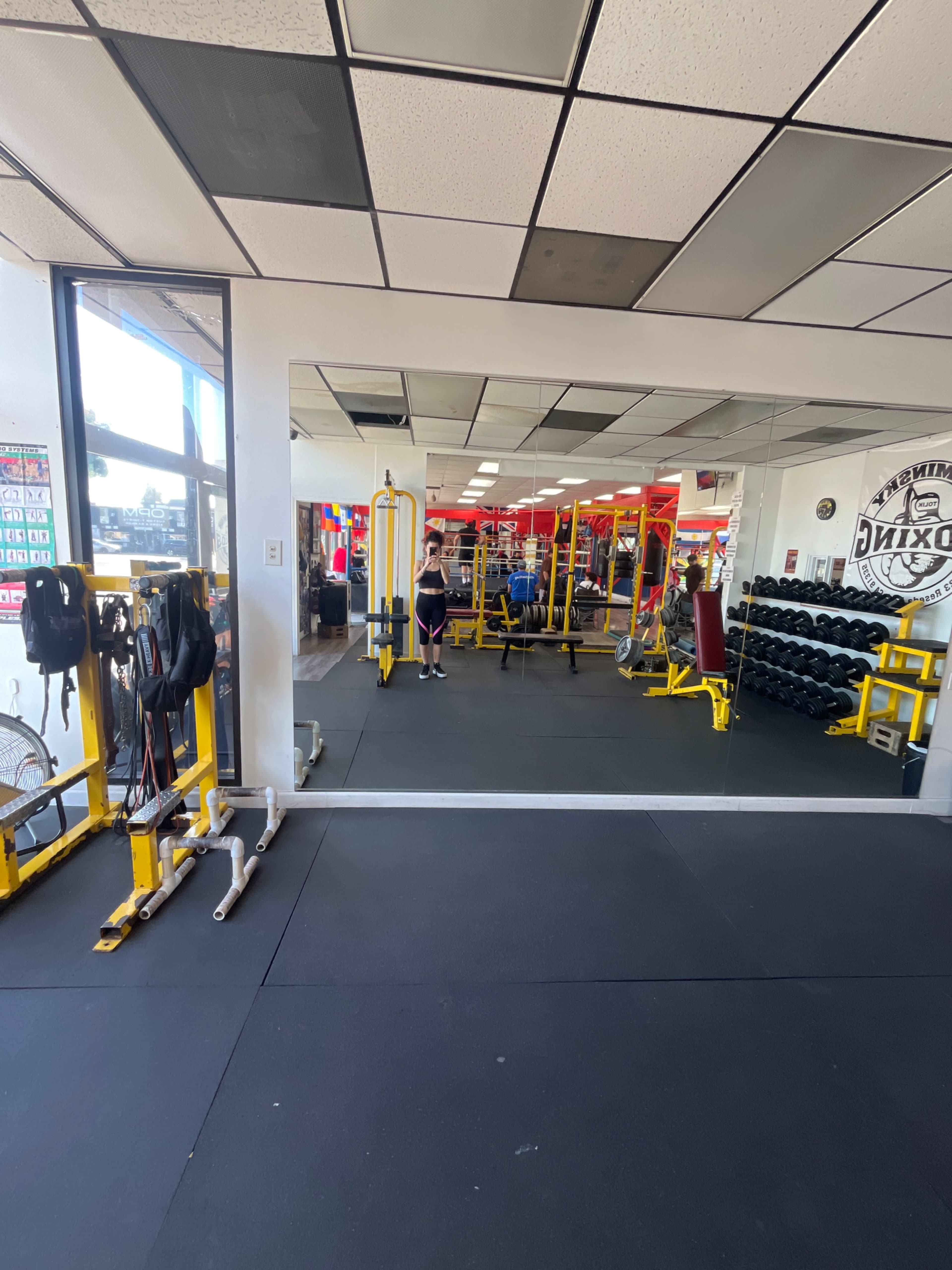 The image shows a gym interior with yellow weightlifting equipment, dumbbells on racks, and a large mirror reflecting part of the space.
