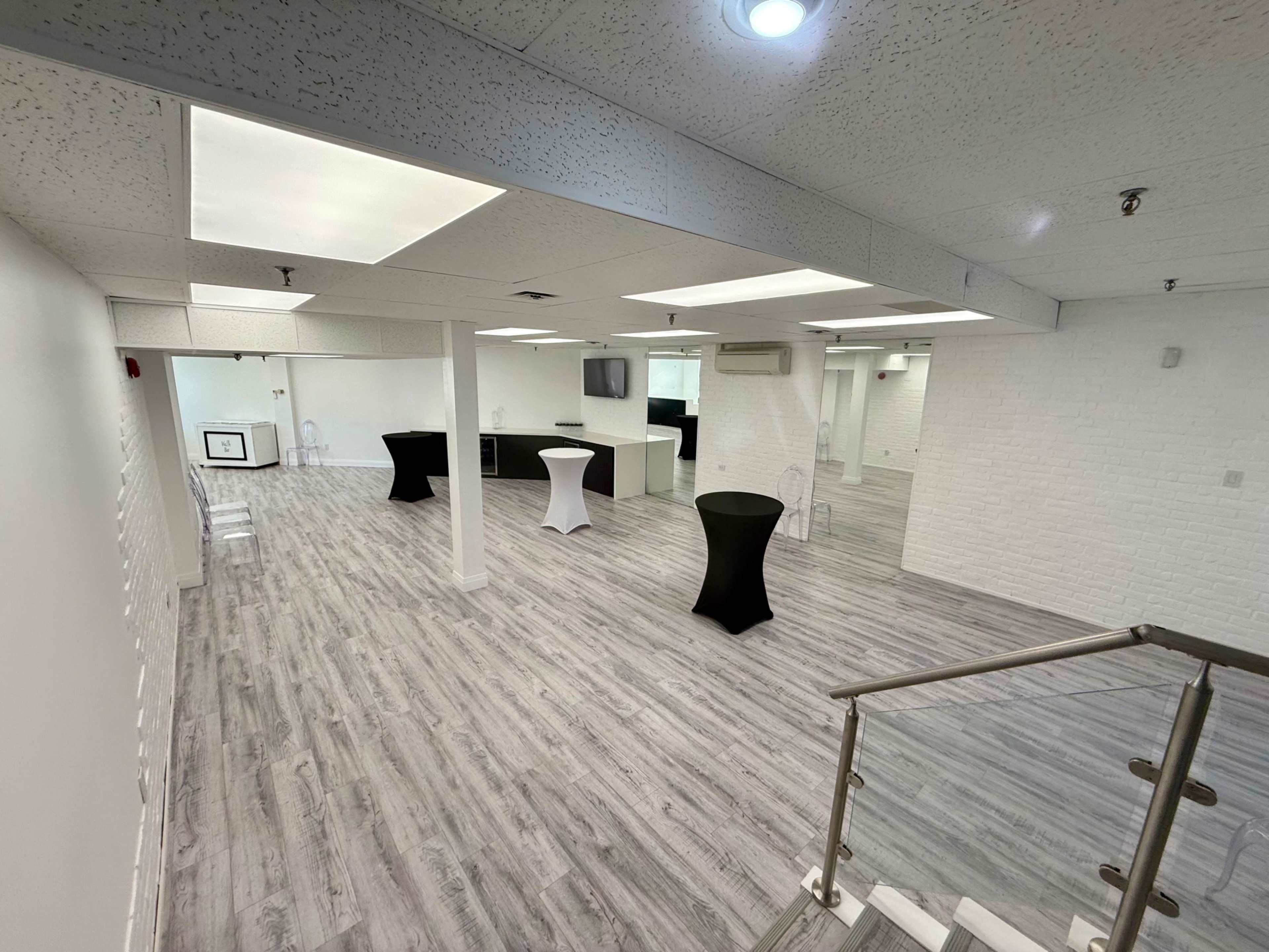 The image shows a spacious and modern room with light-colored flooring, white walls, and a bar area, featuring several high-top tables.
