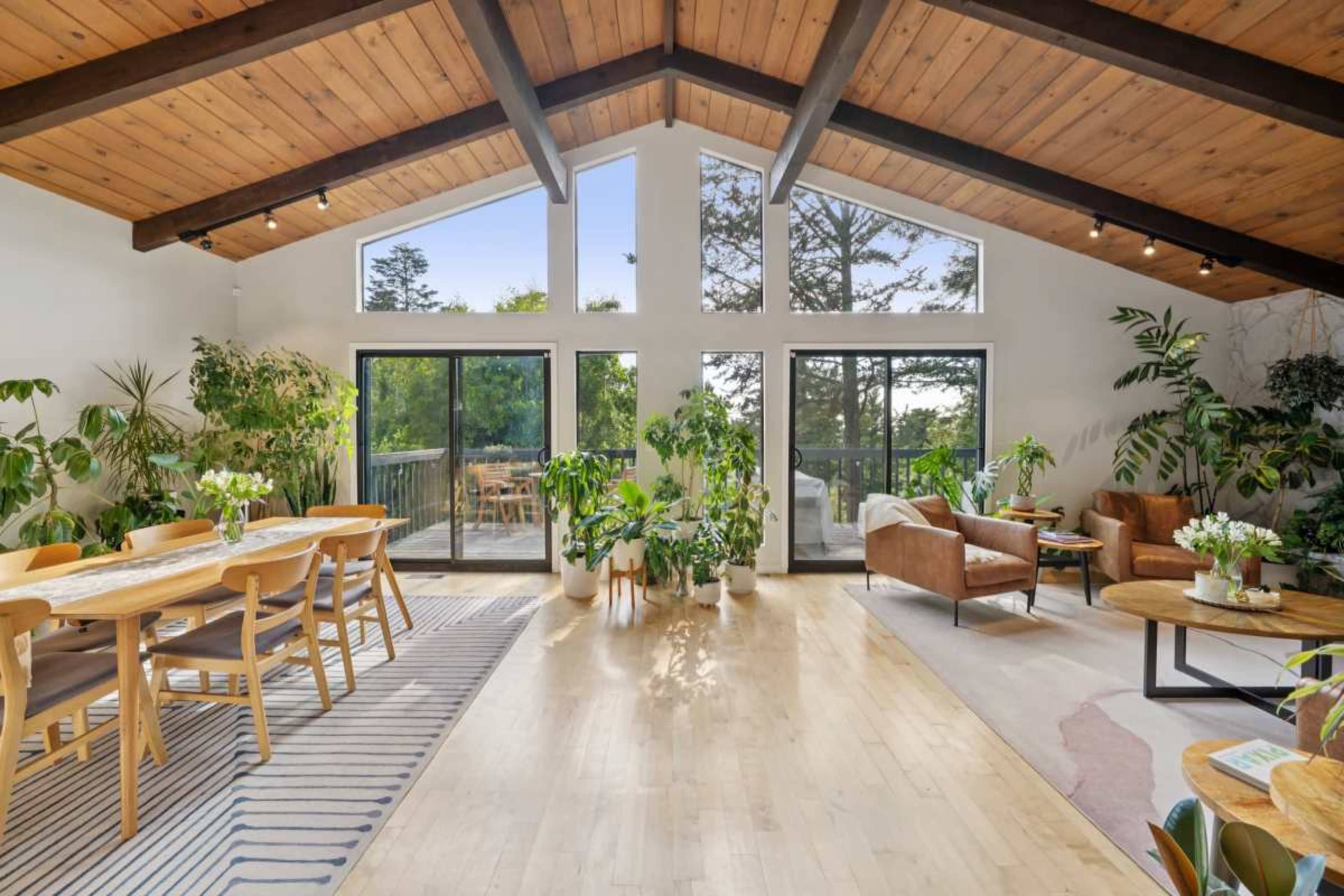 The image shows a spacious, well-lit living area featuring a dining table, comfortable seating, and large windows overlooking greenery.
