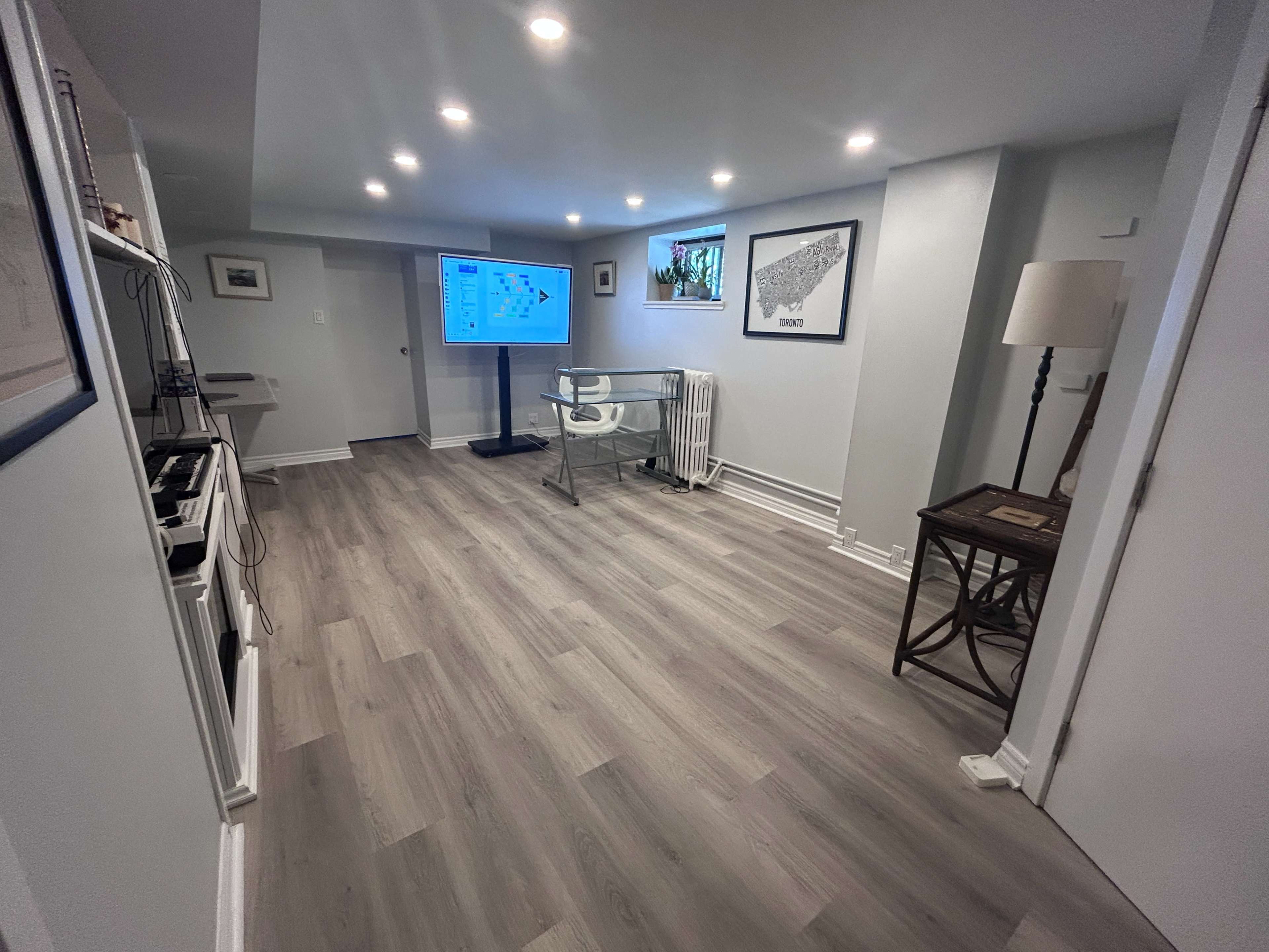 The image shows a well-lit basement room featuring a large screen on a stand, a chair, and wooden flooring, along with framed pictures on the walls.