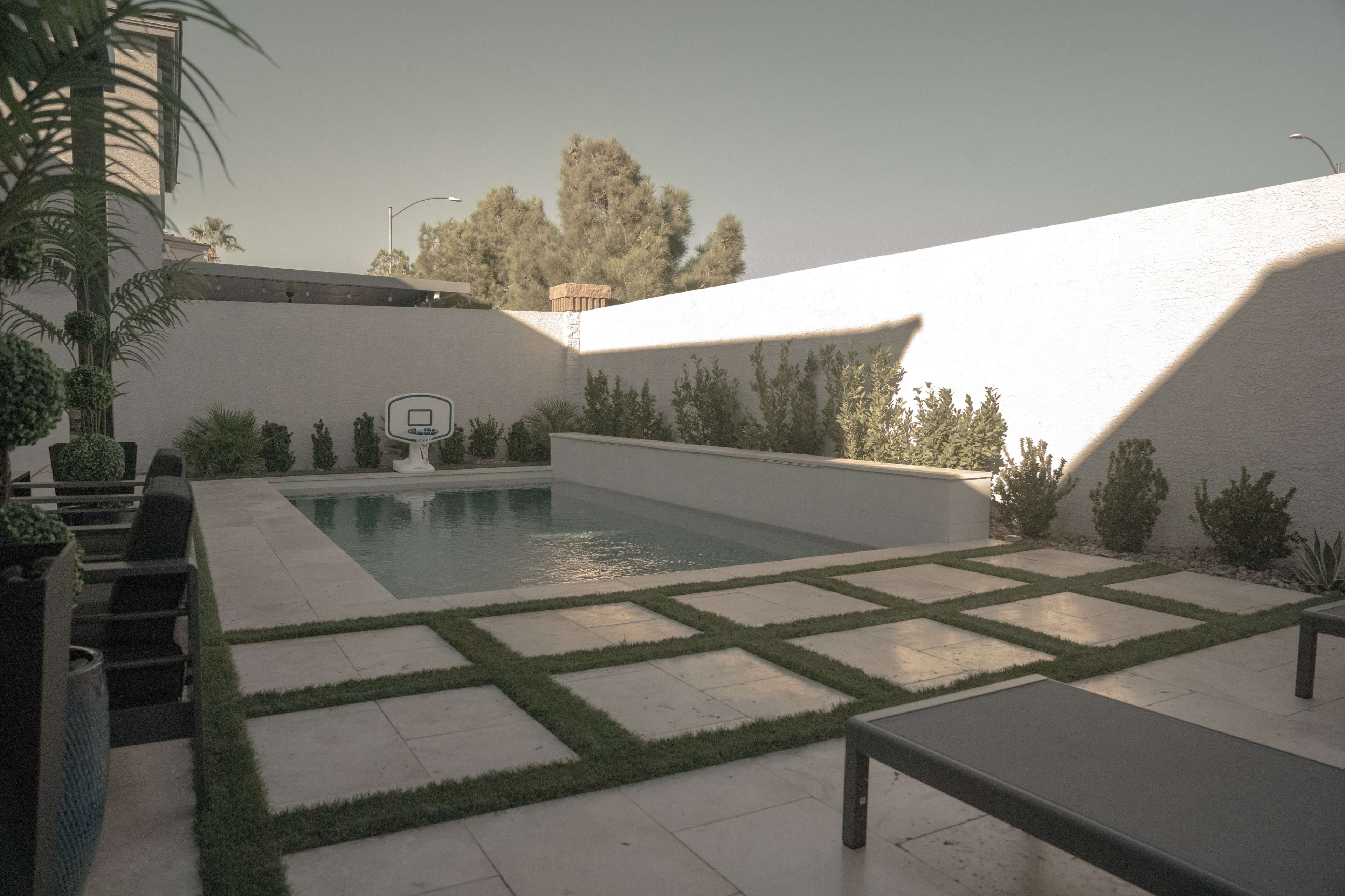The scene features a modern backyard with a rectangular swimming pool surrounded by concrete pavers and grassy areas, with landscaped plants against a white wall.