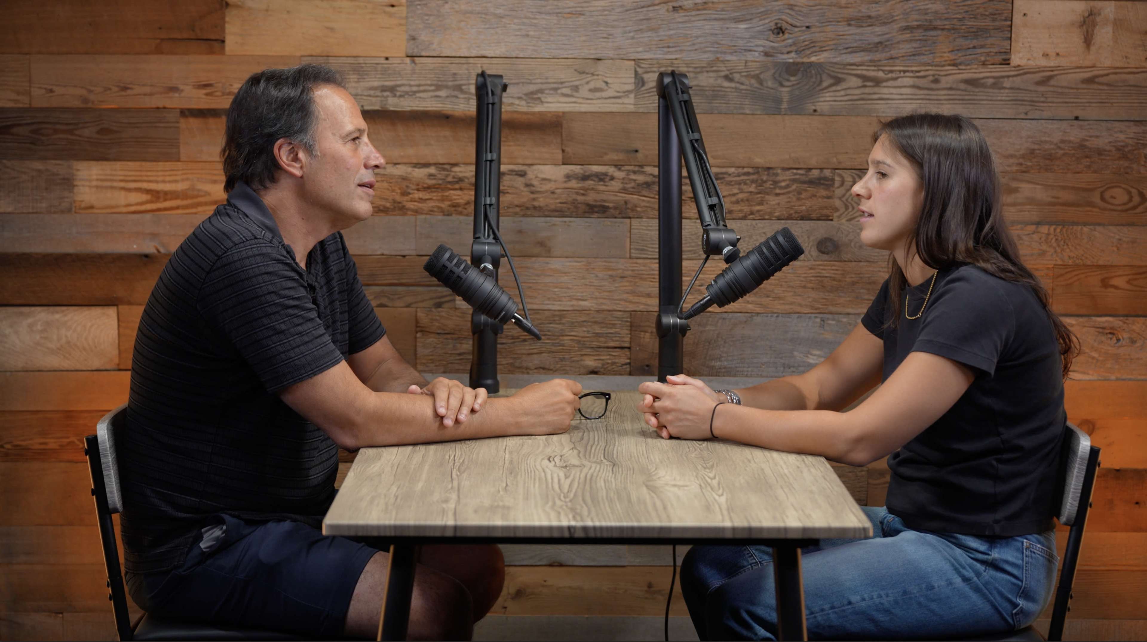 A man and a woman are sitting at a table with microphones in front of them, engaged in a conversation against a wooden wall backdrop.