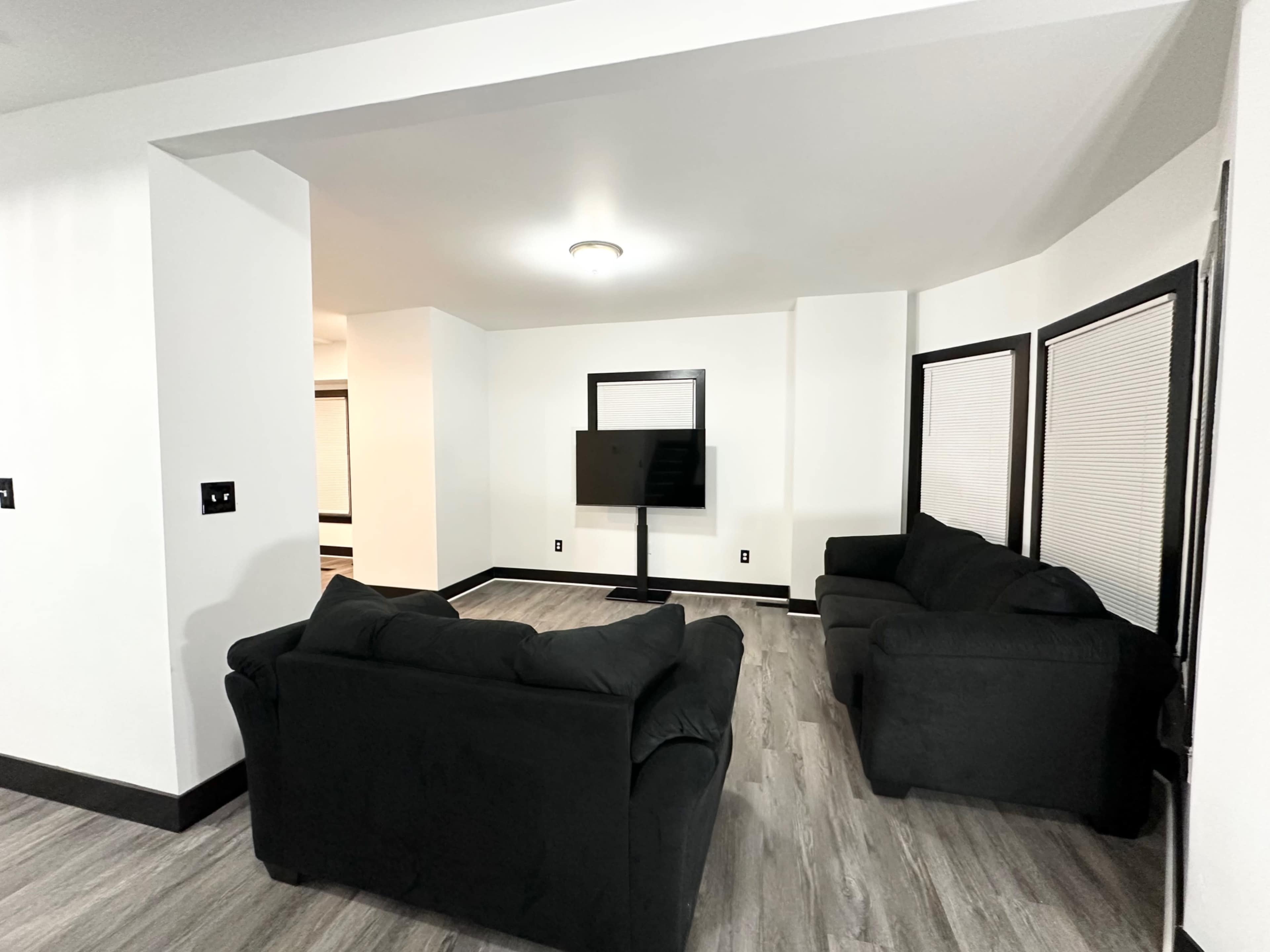 A modern living room features two dark sofas facing a television on a stand, with large windows partially covered by blinds.