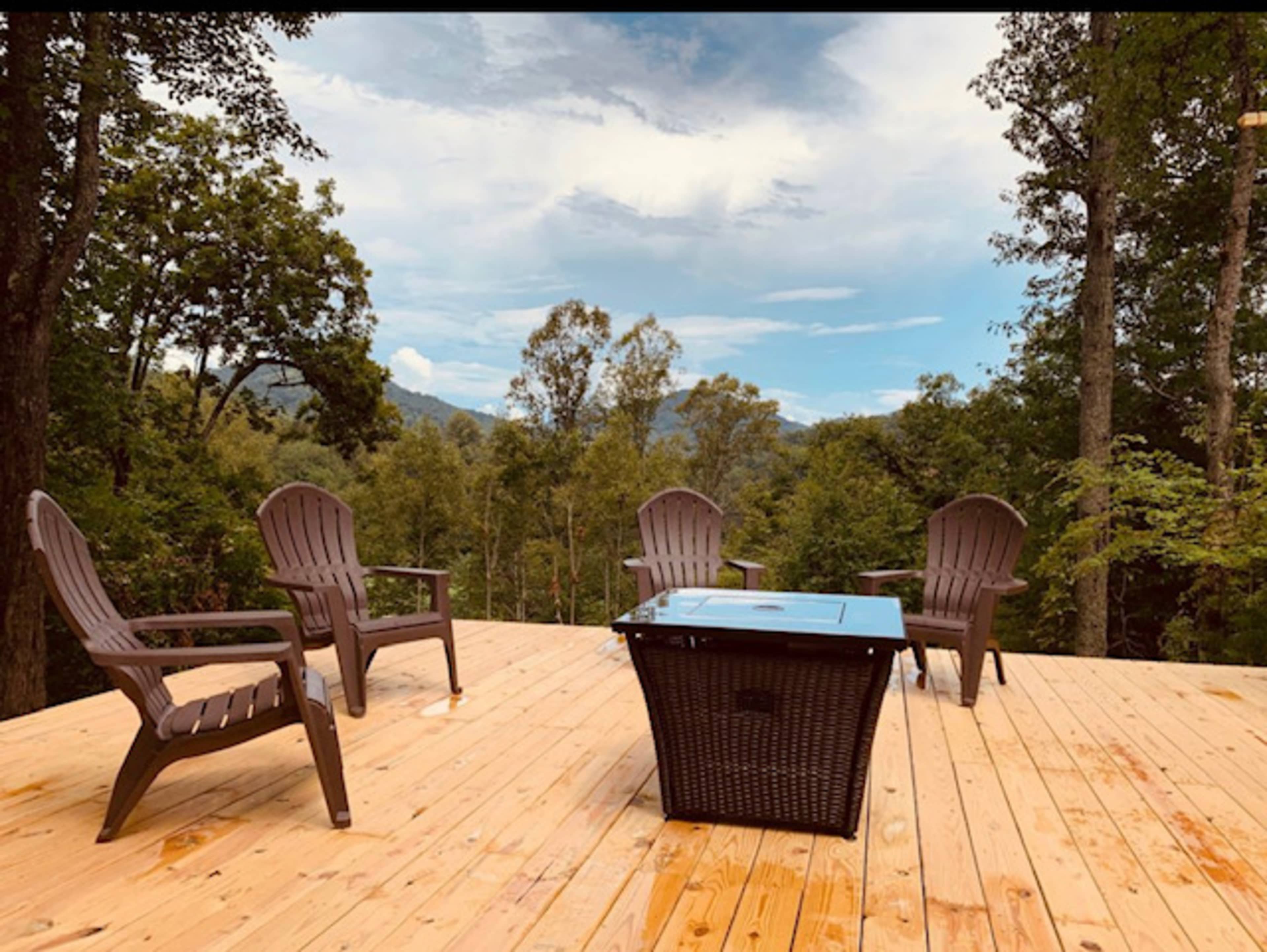 A wooden deck featuring four brown Adirondack chairs and a square coffee table surrounded by trees and mountains in the background.