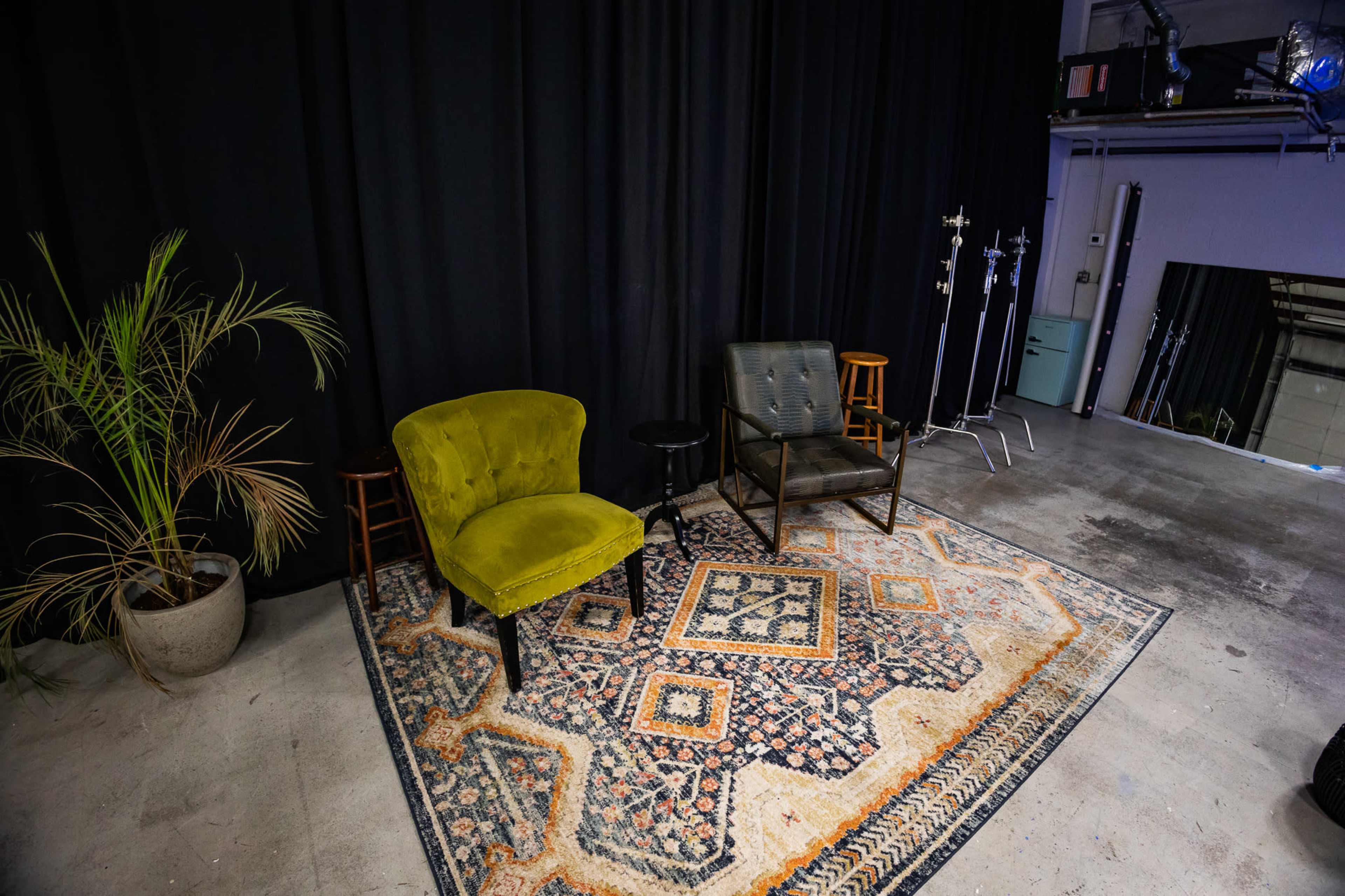 The image shows a small studio area with two armchairs, a side table, and a patterned rug, accompanied by a potted plant against a black backdrop.