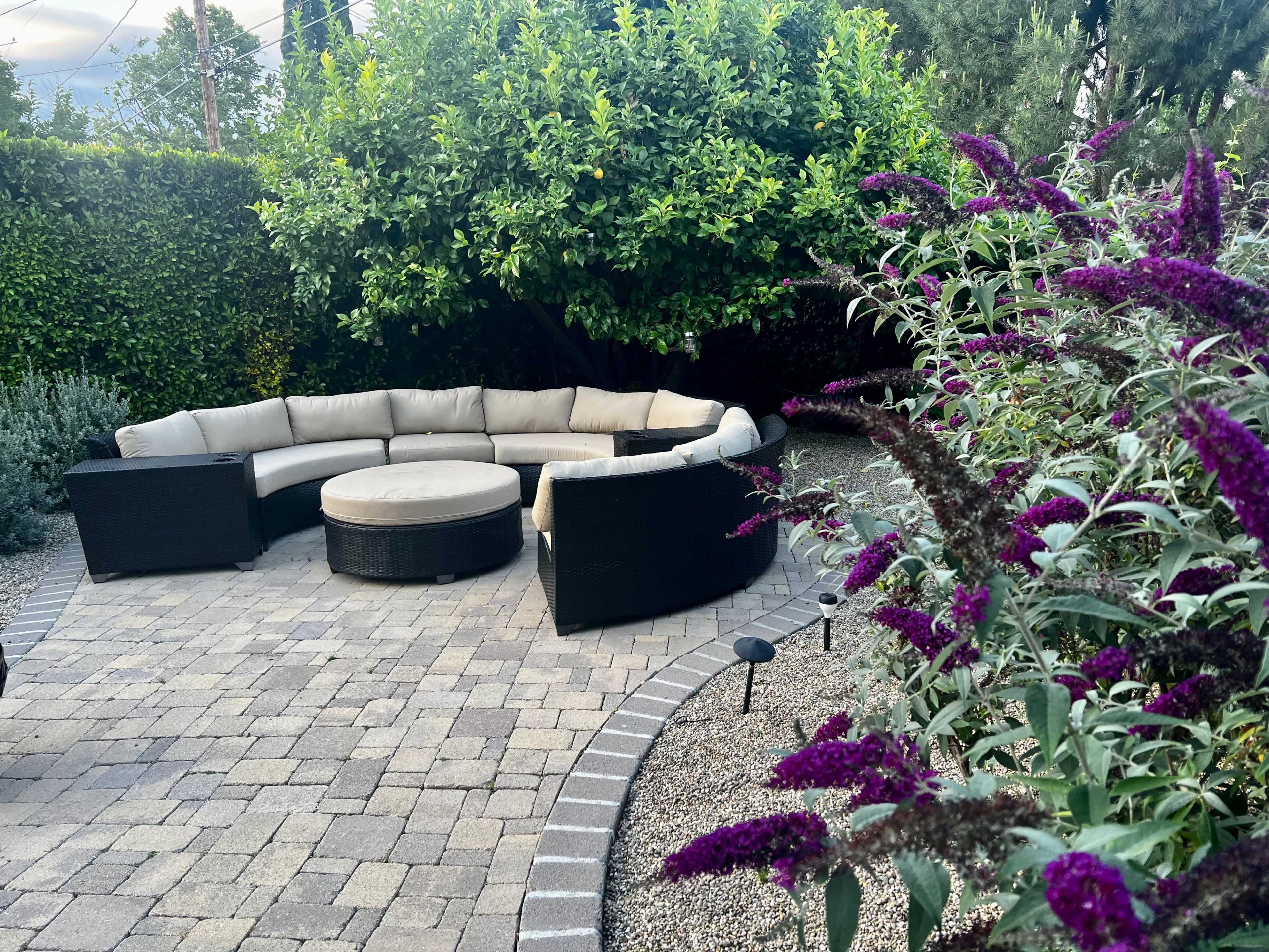 A circular seating area with a beige cushion sofa is surrounded by lush greenery and purple flowering plants on a stone pathway.