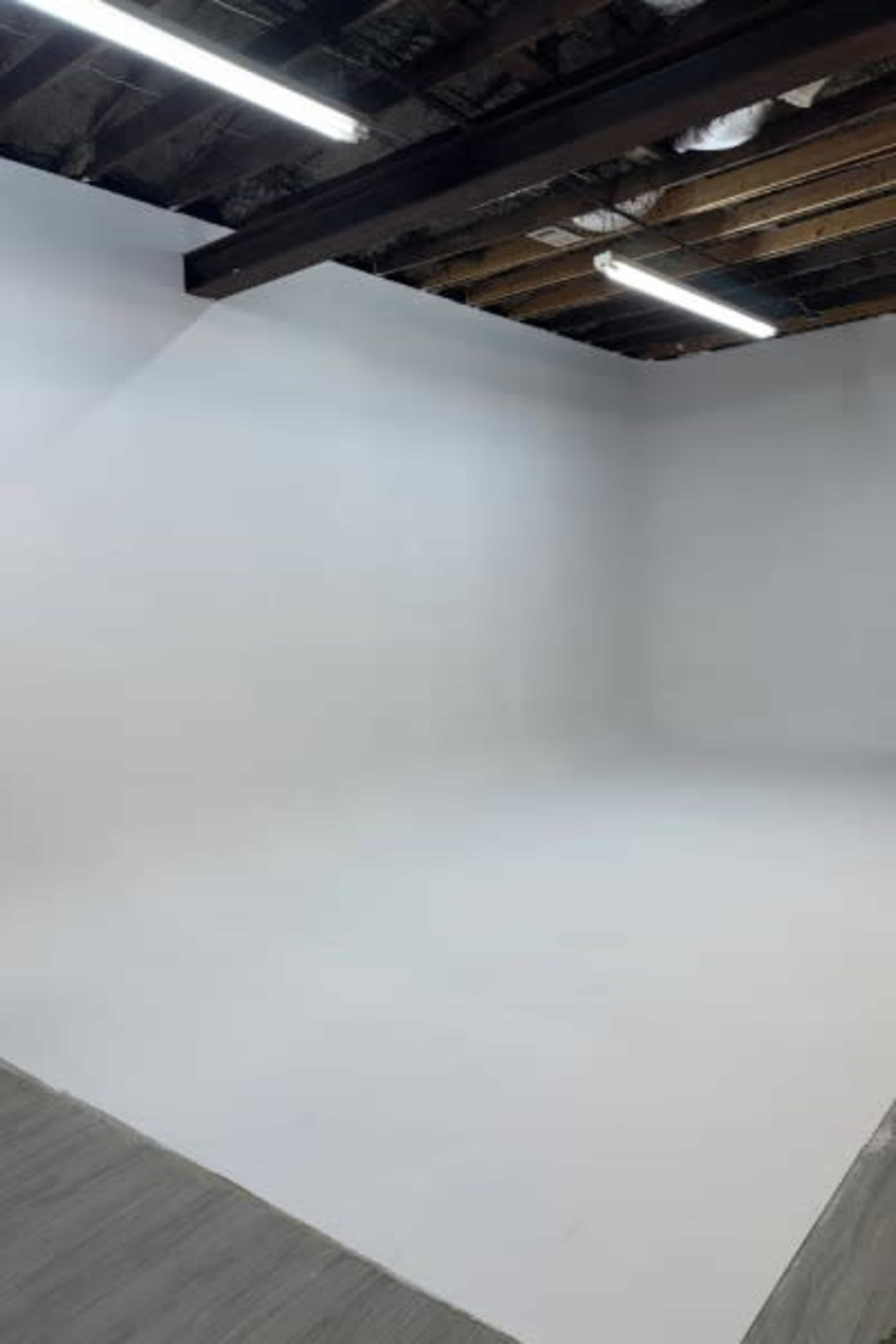 The image shows a minimalistic studio space with white walls and exposed wooden beams overhead.